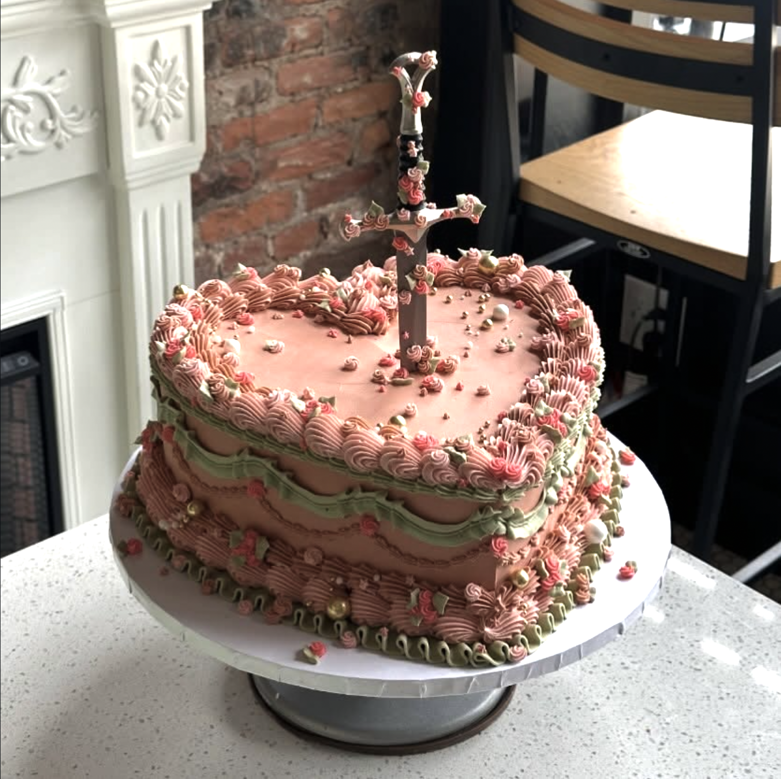 A heart-shaped pink birthday cake decorated with pink and green frosting flowers, gold-colored balls, and a sword-shaped cake topper, placed on a white cake stand on a white speckled countertop.