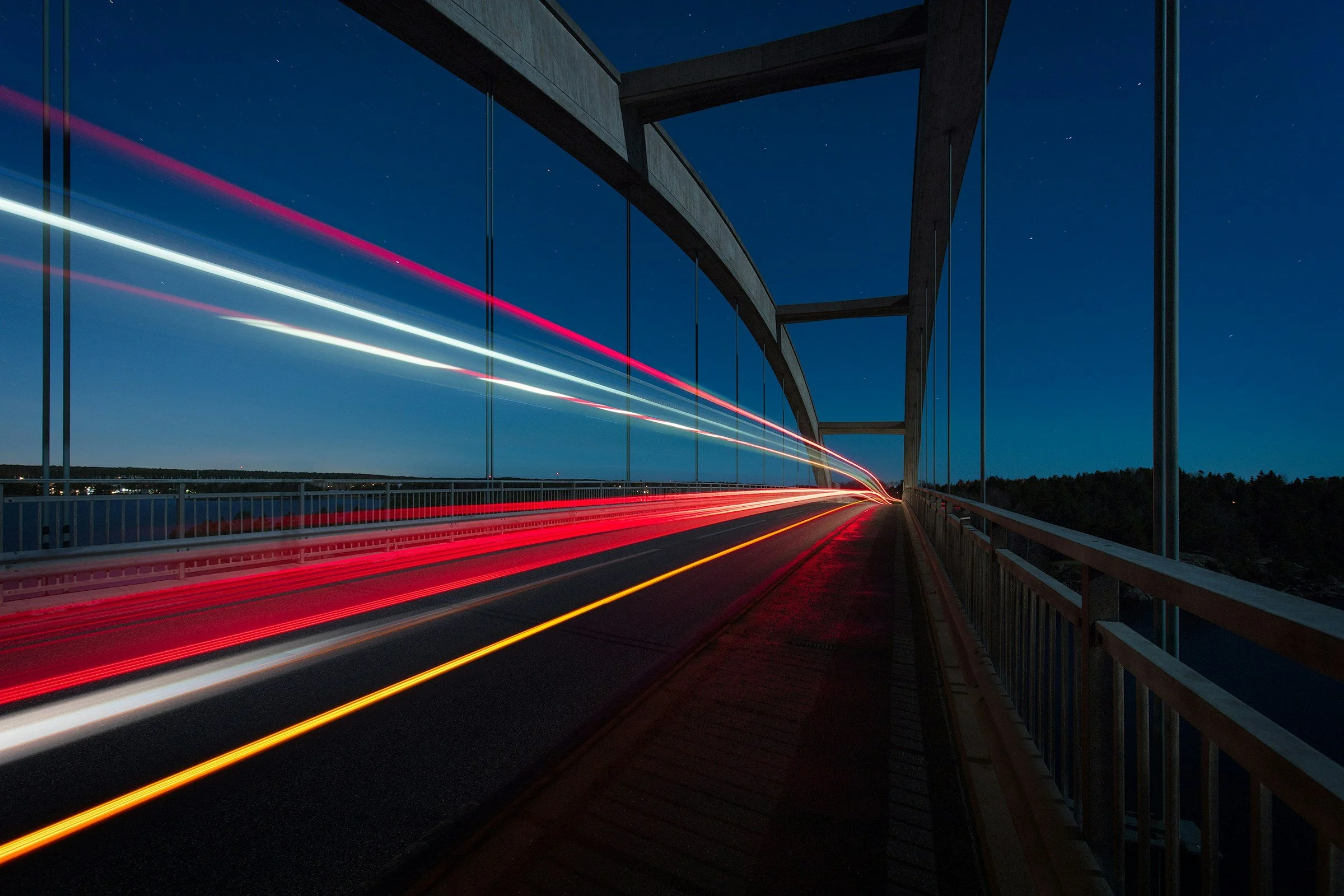 Nighttime photo of a bridge with car light trails creating streaks of red, white, and yellow, with a dark blue sky and stars in the background.