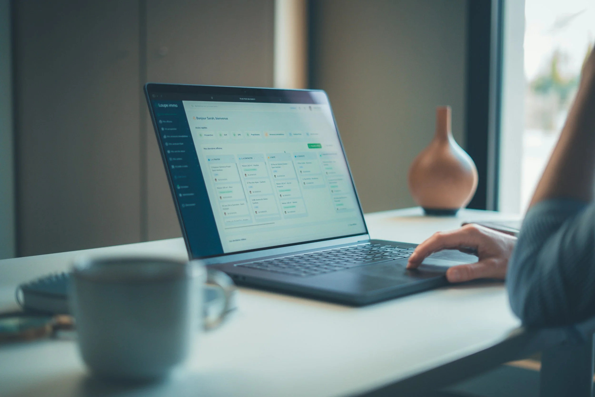 A person working on a laptop displaying a project management or task organization software on a white desk, with a white coffee mug and a decorative vase in the background near a window.