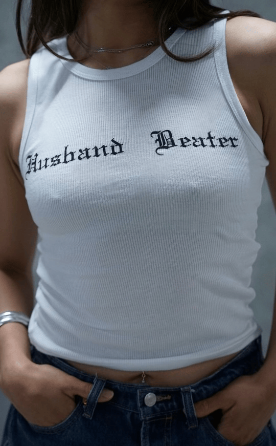 A woman wearing a white ribbed tank top with the words "Husband Beater" printed on it, and blue jeans.