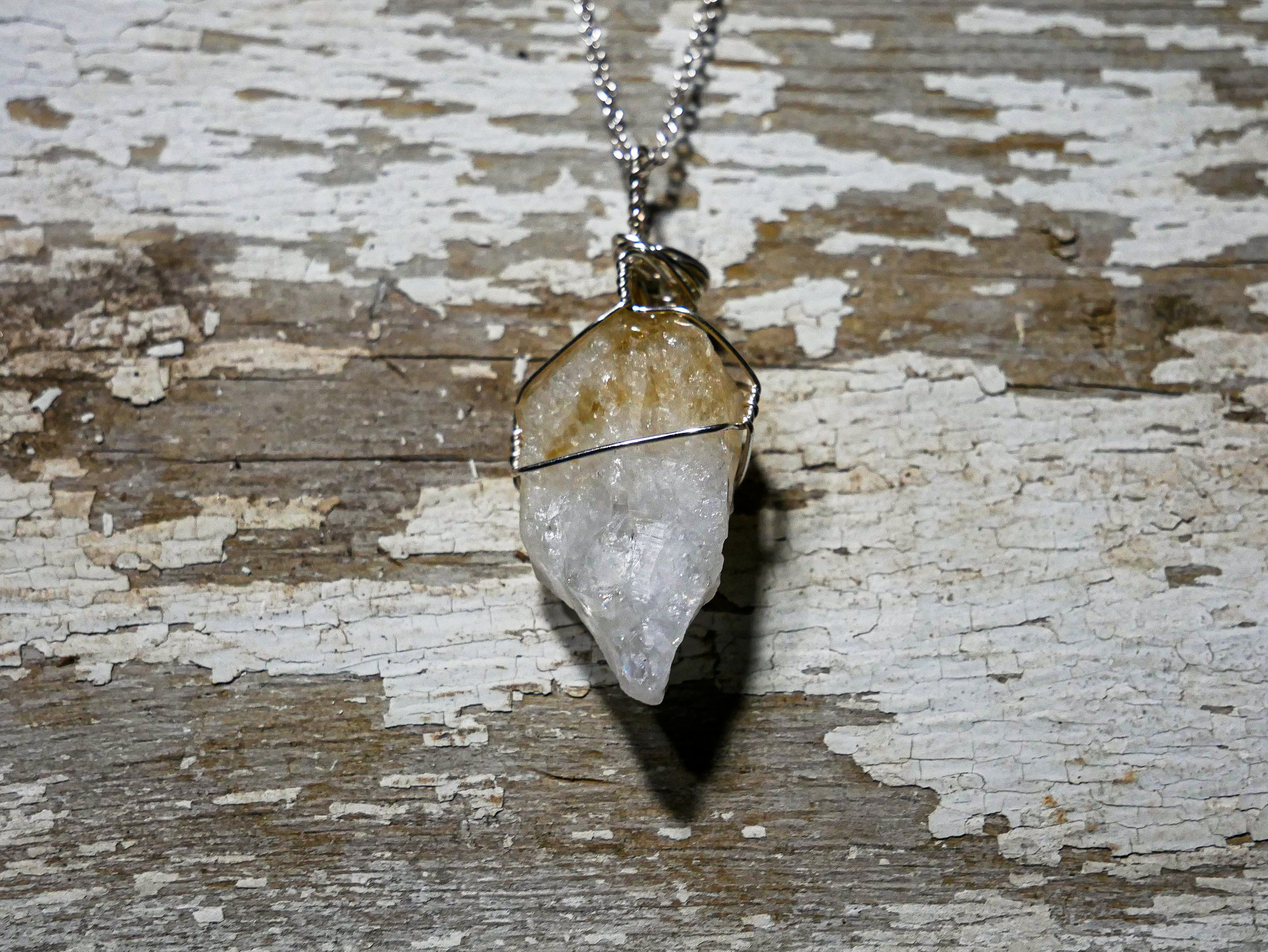 A pendant necklace with a raw crystal stone wrapped in wire, hanging from a silver chain, on a weathered wooden surface.