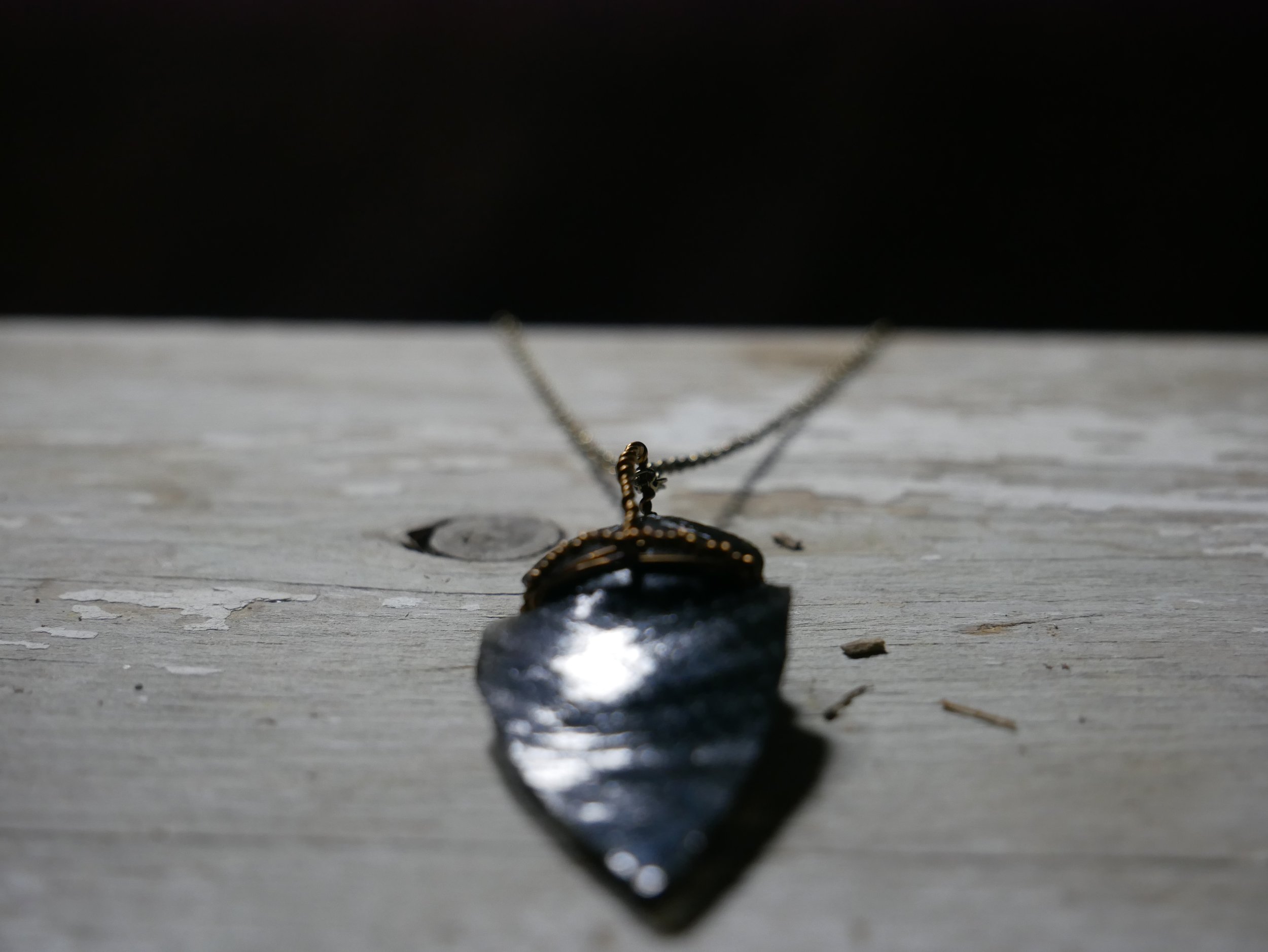 A snake-shaped necklace resting on a weathered wooden surface with a large reflective arrowhead pendant hanging from a chain.