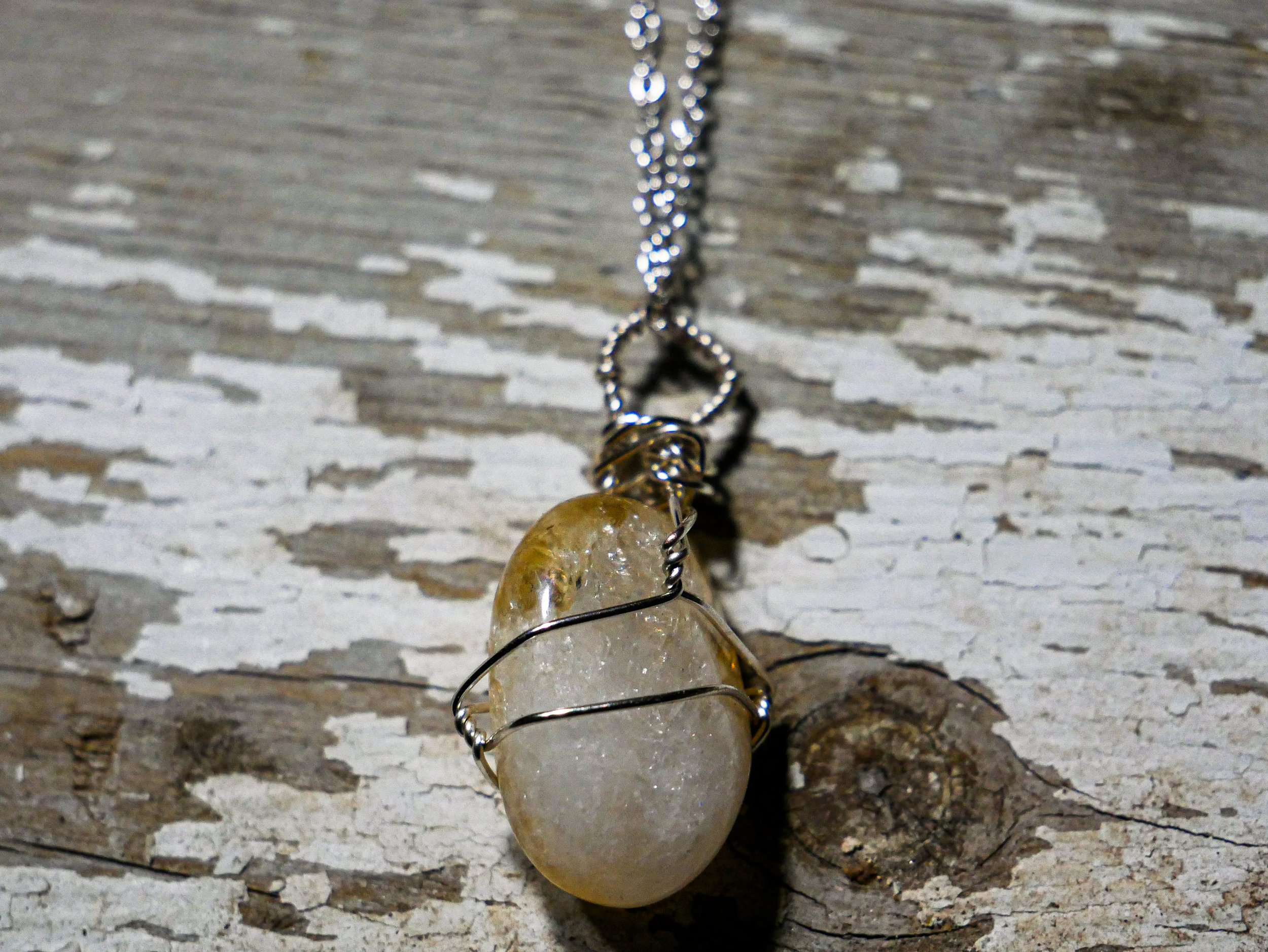A pendant necklace with a yellowish, semi-transparent stone wrapped in wire, hanging from a silver chain against a weathered wooden background.