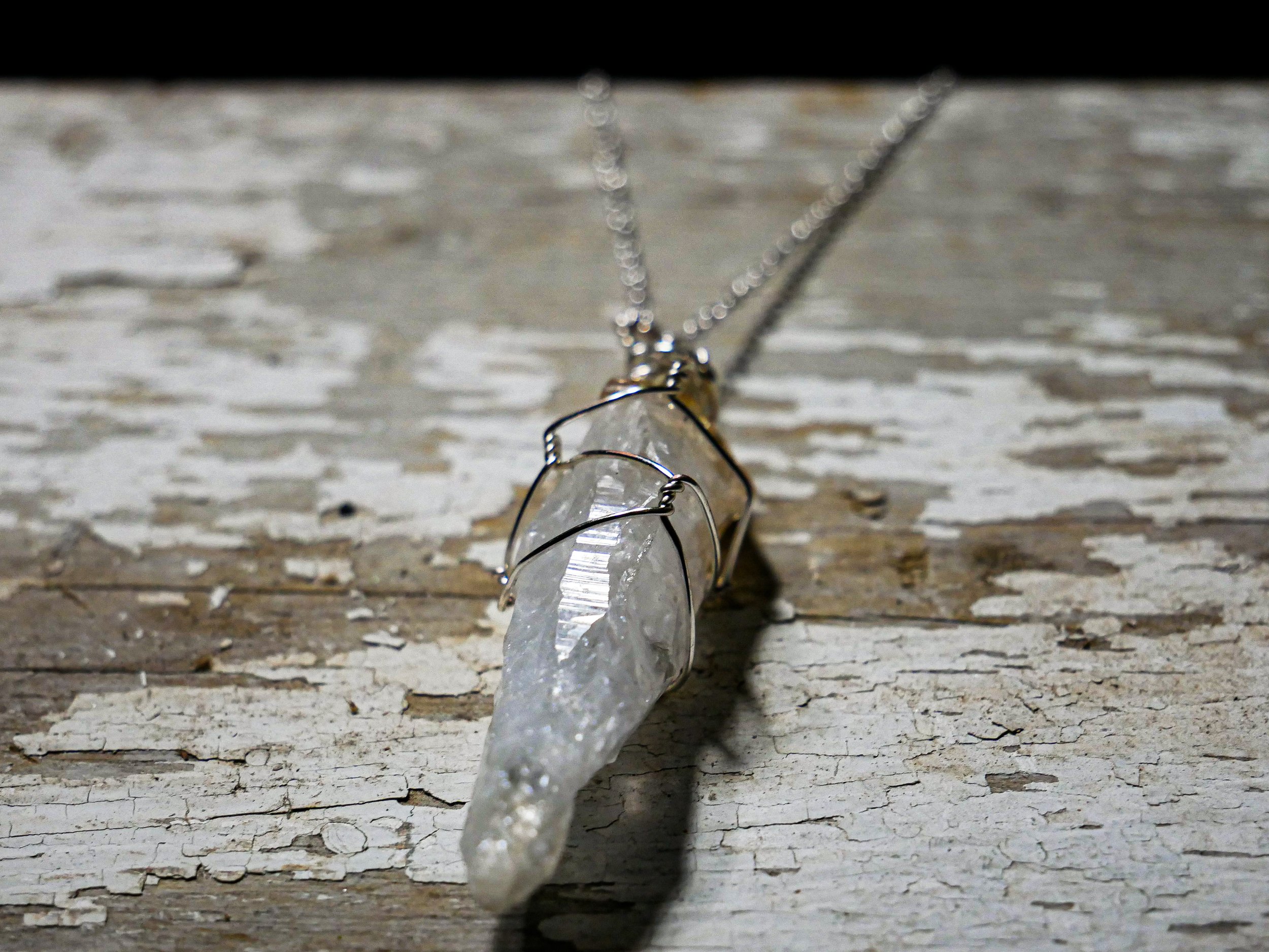 A necklace with a clear, irregularly shaped crystal pendant wrapped in metal wire, lying on a weathered, peeling wooden surface.