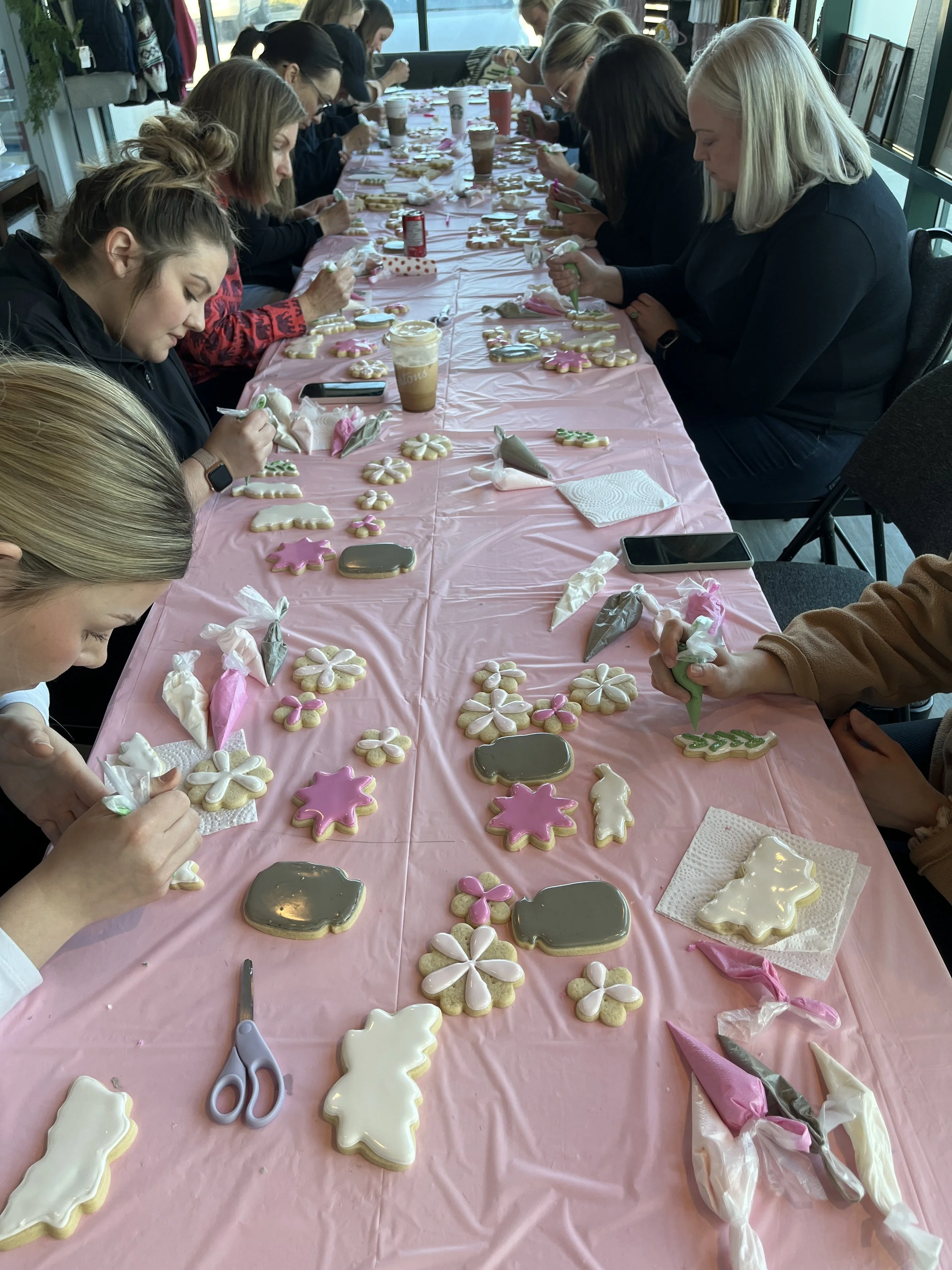 Group of women decorating cookies with pink, white, and green icing on a long pink table covered with a pink tablecloth.
