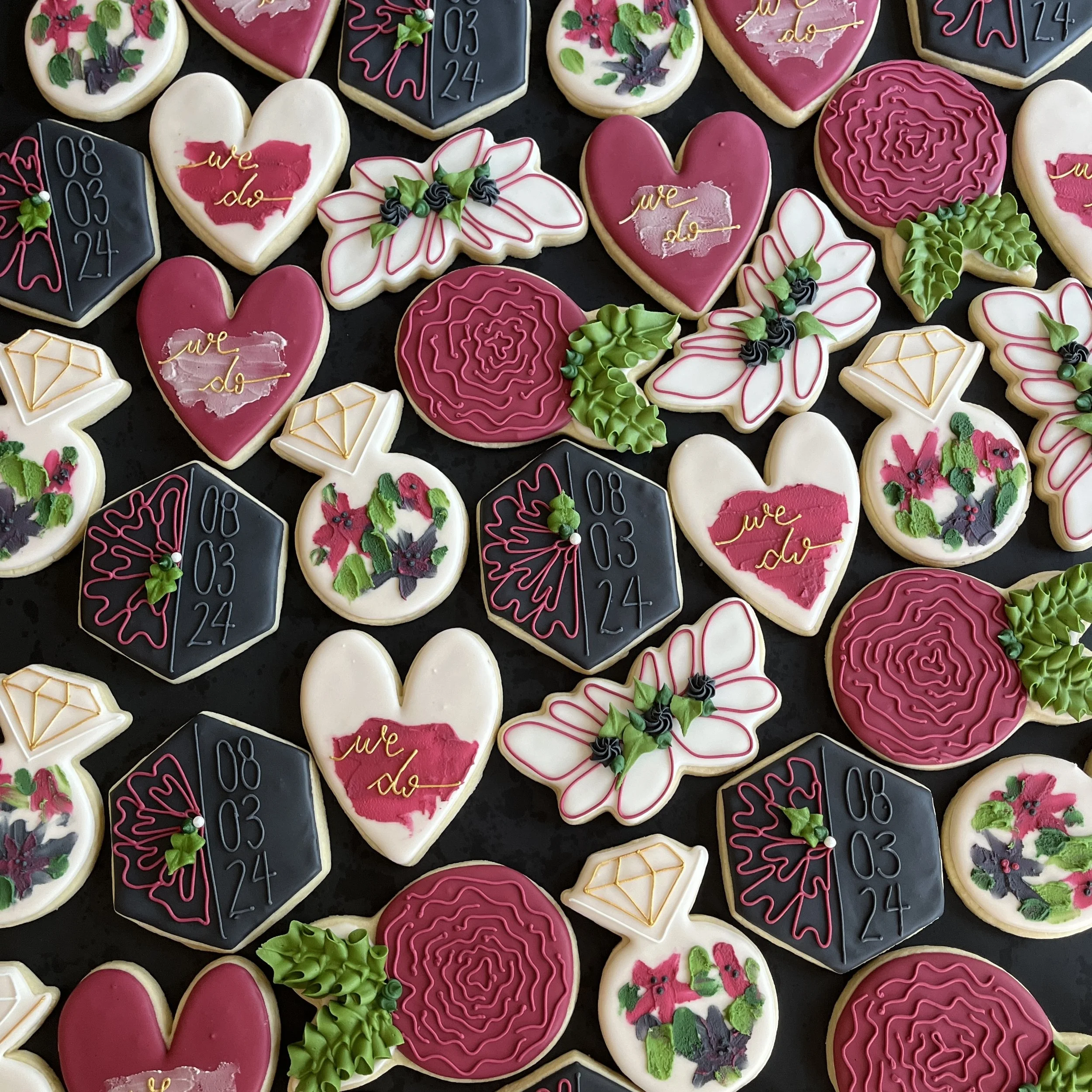 Assorted decorated sugar cookies featuring holiday themes with pink, white, black, and green icing. Designs include hearts, flowers, holly, diamonds, and date-themed cookies with the date "08.03.24." Some cookies have the phrase "we do" written in gold icing, while others have festive and floral decorations.