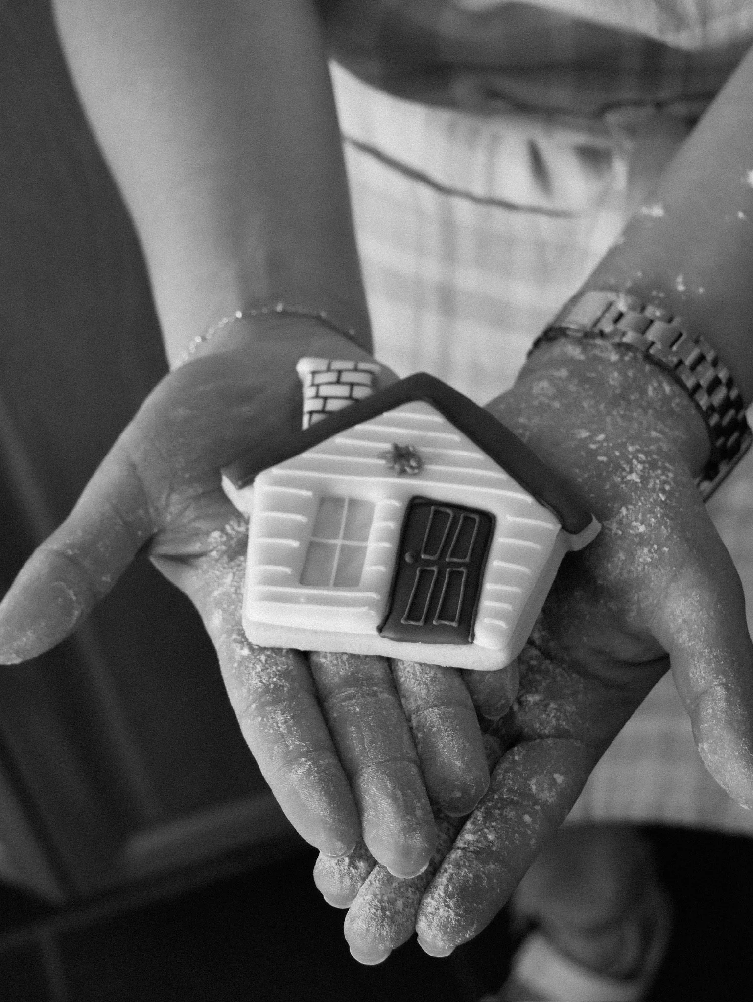 A person's hands holding a small plastic house toy with a black door and window. The hands are covered in white dust or powder, and the person is wearing a wristwatch and a bracelet.