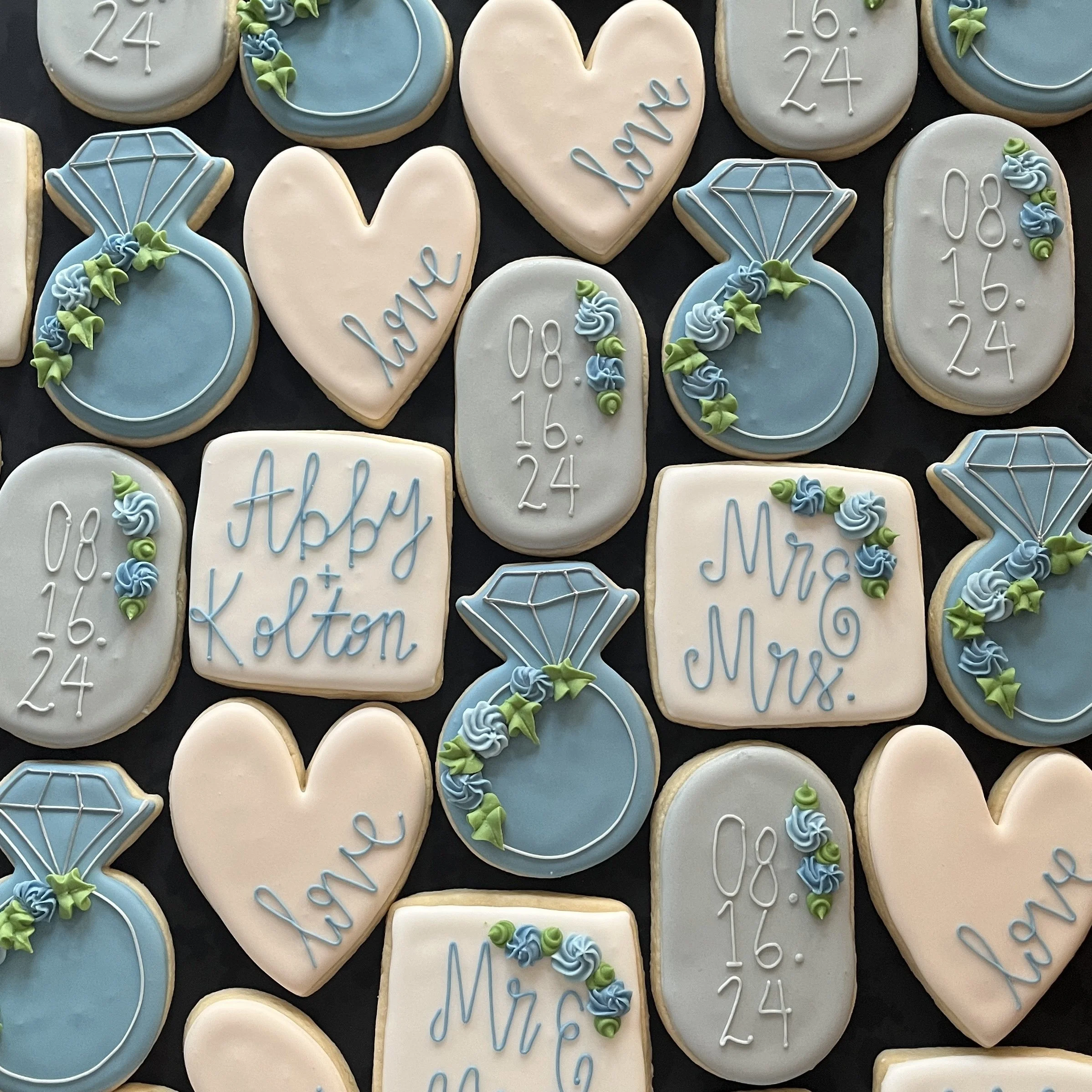 Decorated cookies for a wedding or anniversary, featuring messages like "love," "Mr. & Mrs.," and "Abby & Kolton," along with date "08.16.24" and diamond ring shapes, all in pastel colors.