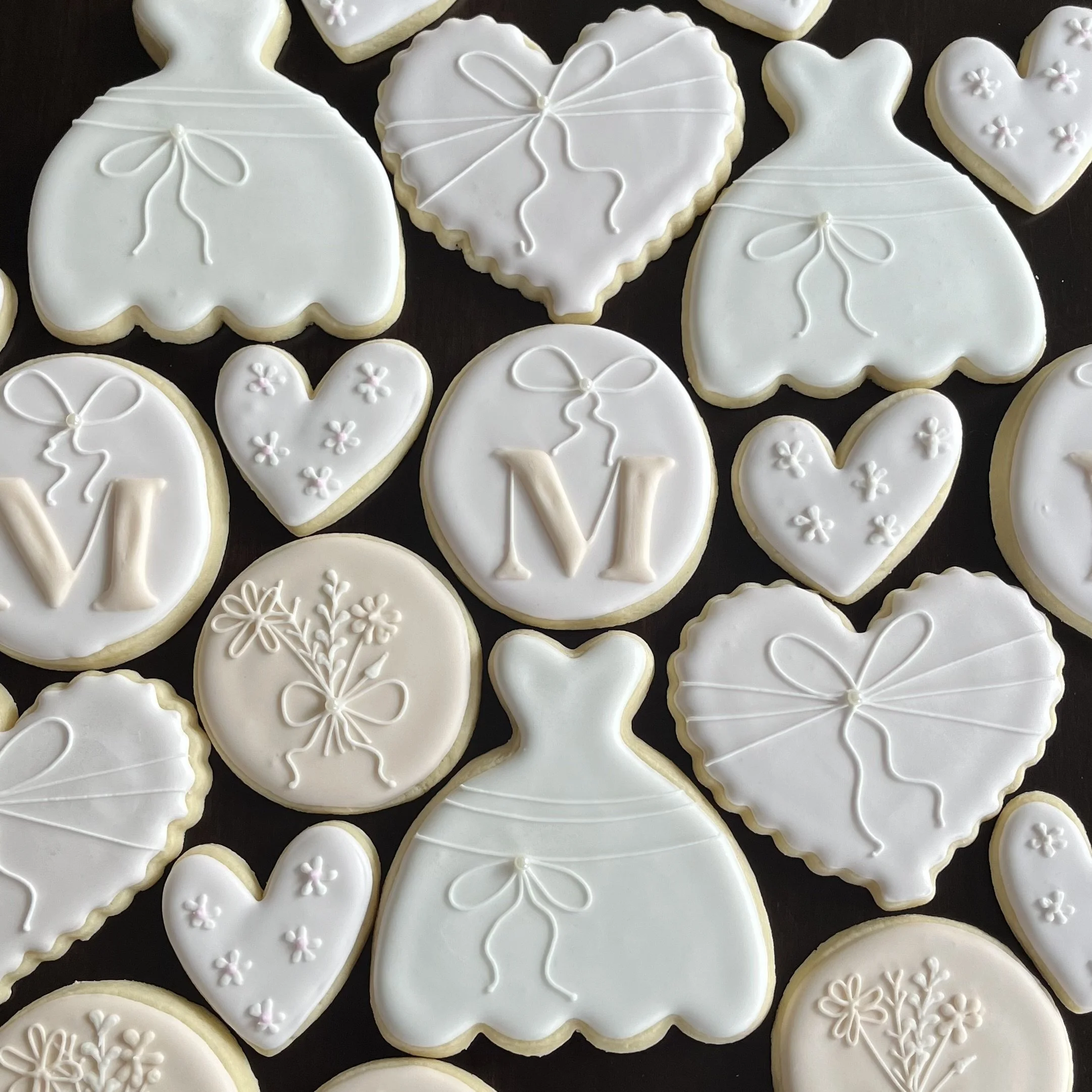 Assorted decorated cookies with white icing, featuring heart-shaped and dress-shaped designs, decorated with bows, flowers, and the letter M.