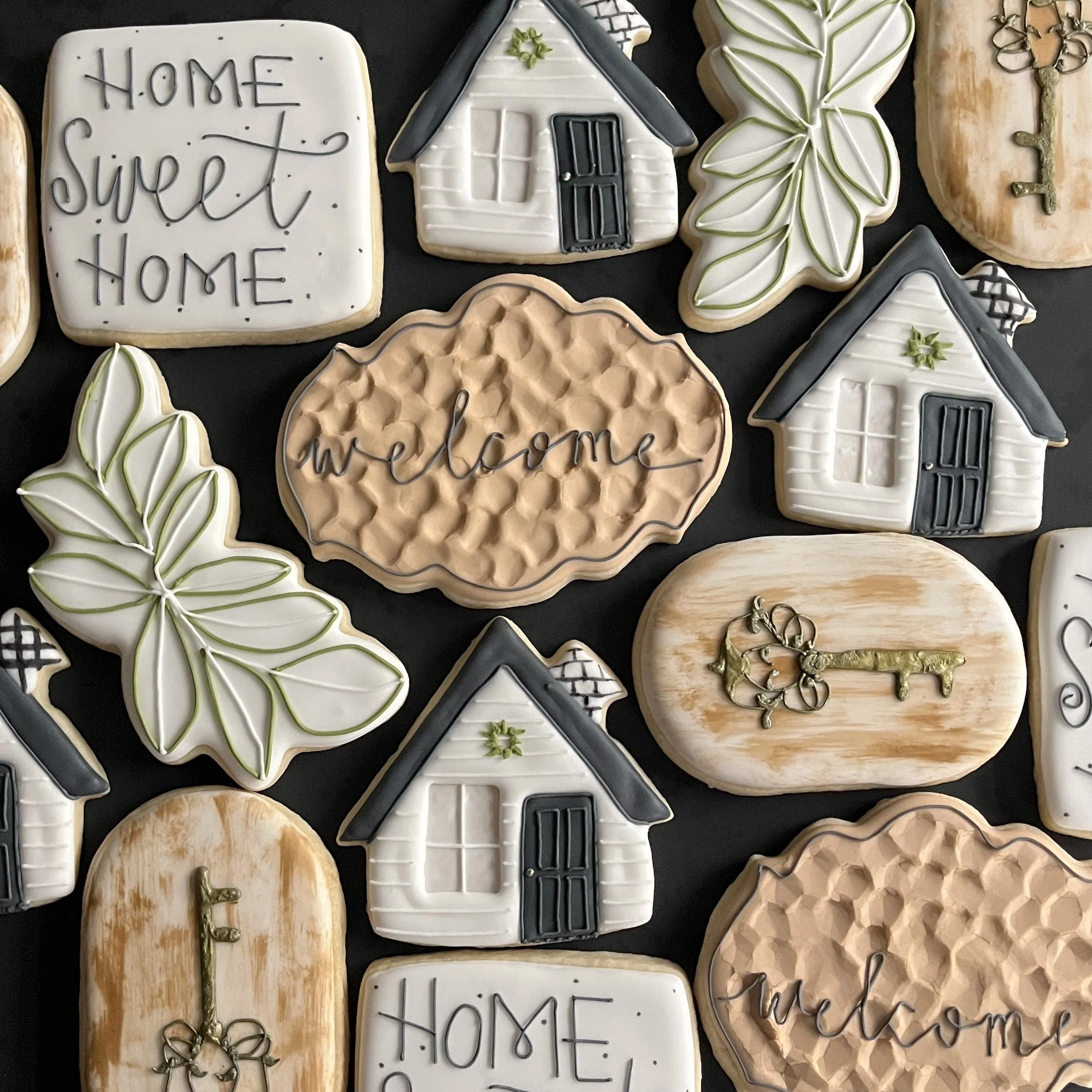 Decorative cookies with home and welcoming themes, shaped like houses, leaves, and keys with white, black, and gold icing.