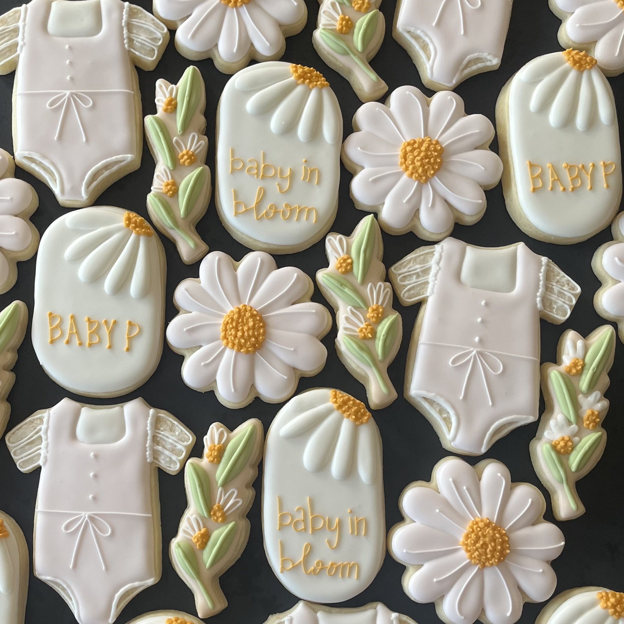 Decorative cookies shaped like daisies, baby onesies, and floral designs with white, yellow, and green icing, some with phrases like "baby in bloom" and "BABY" in yellow icing.