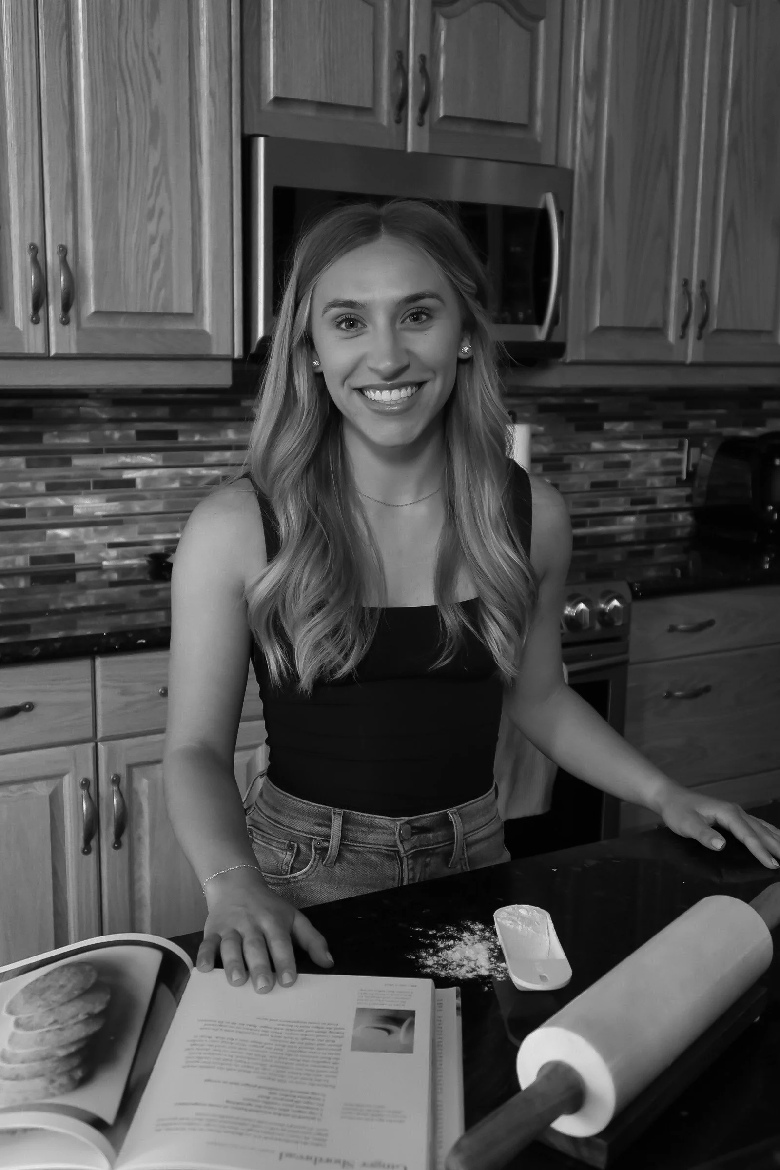 A young woman with long wavy hair smiling in a kitchen, standing behind a counter with a rolling pin, a recipe book, and a scoop of flour.