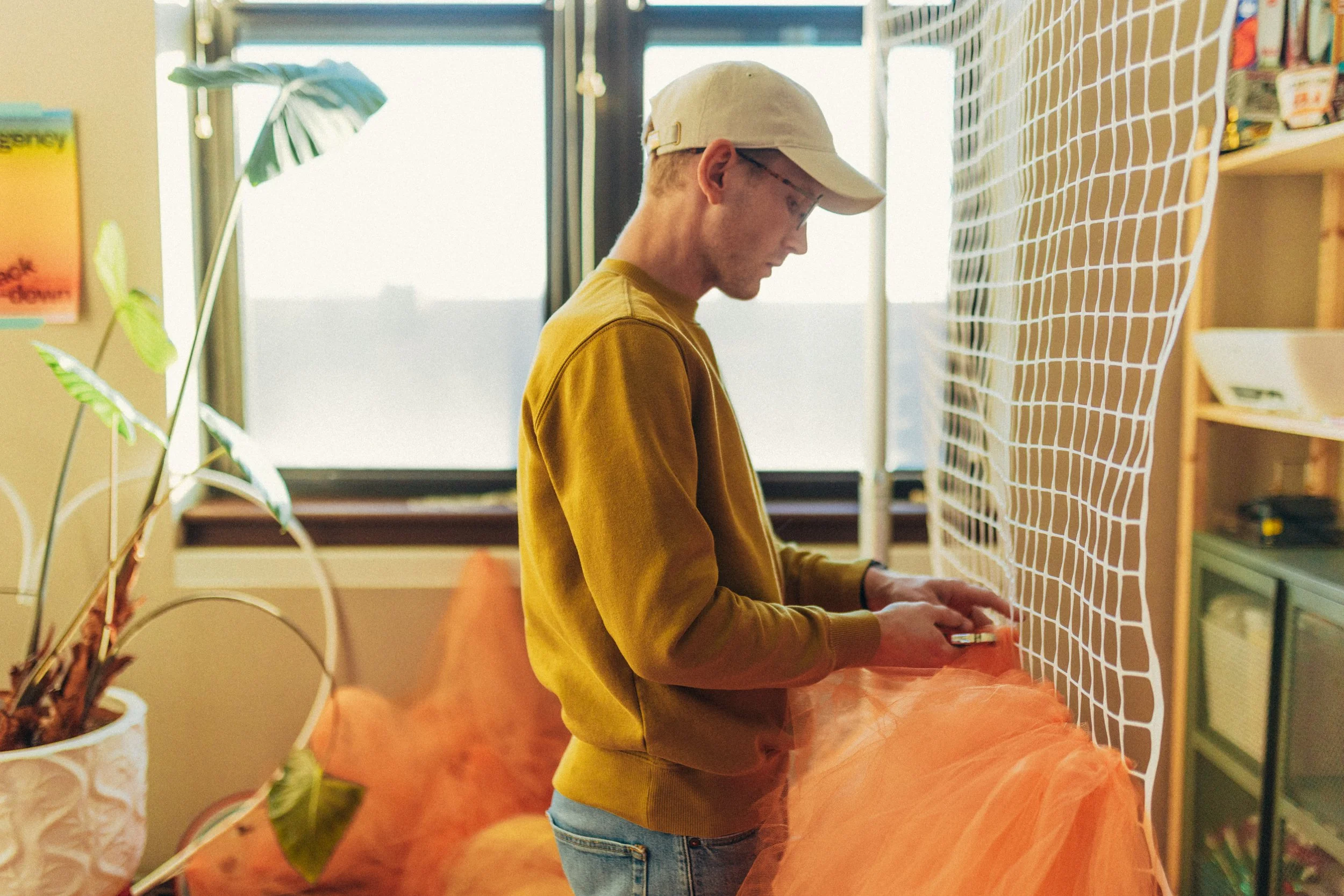 A young man in a yellow sweatshirt and white cap looking at his phone while standing in a room with a large window, a green plant, and orange curtains.