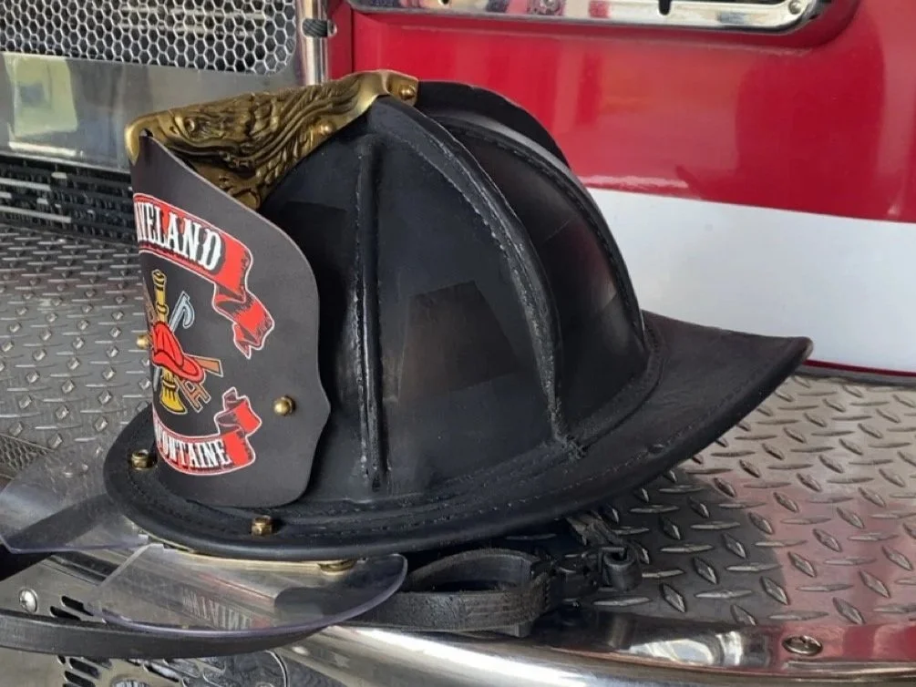 A firefighter helmet with a badge reading 'HIGHLAND VOLUNTEER FIRE DEPARTMENT' resting on a metal surface, with part of a fire truck visible in the background.