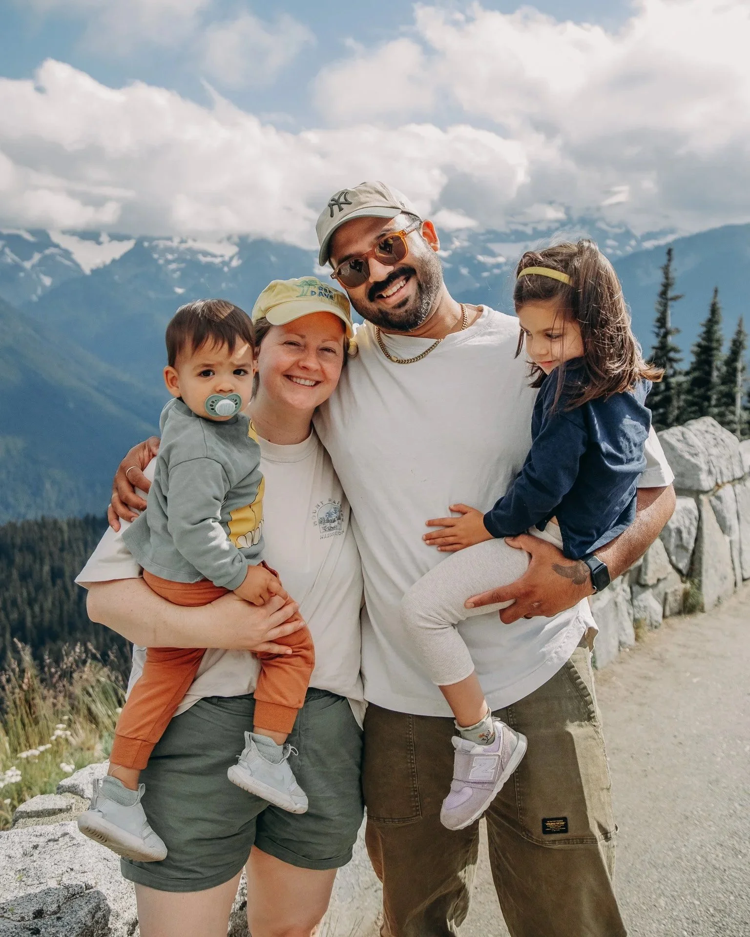 A family of four smiling and hugging outdoors with mountains and trees in the background, during daytime.