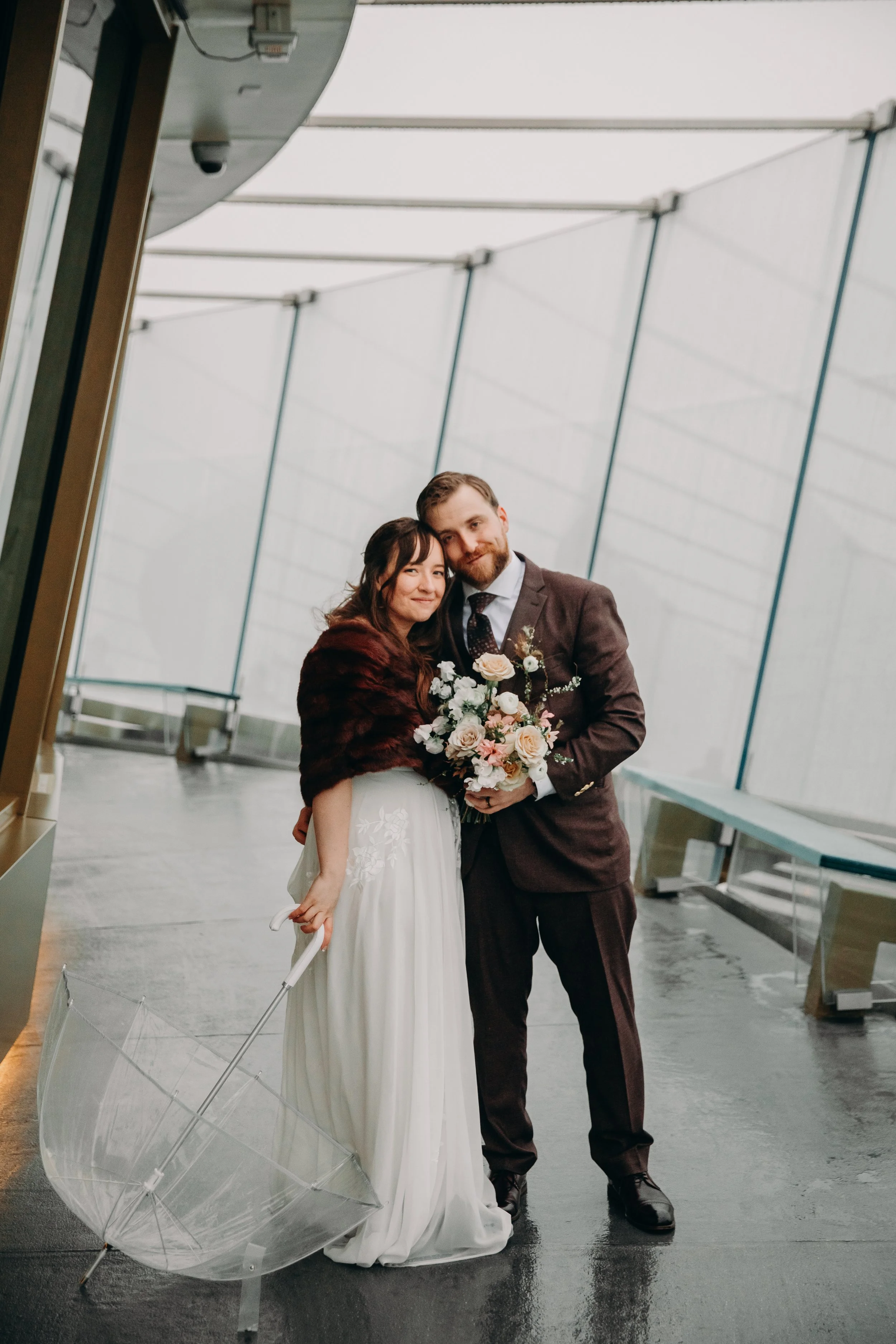 A couple, newlyweds, standing close together indoors, with the woman holding a bouquet of flowers and the man dressed in a suit. The woman has dark hair and is wearing a white dress and a dark-colored shawl or wrap. The man has light brown hair and a beard, wearing a dark suit with a tie. An umbrella is in the foreground, and the background shows a modern, curved architecture with large glass windows.