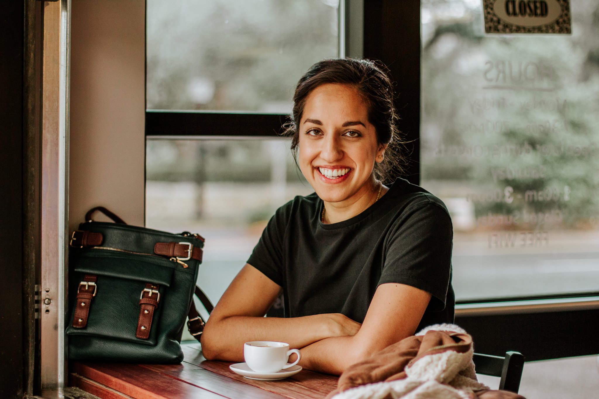 A woman smiling while sitting at a breakfast cafe, with a cup of coffee, a purse, and a cloth on the table.