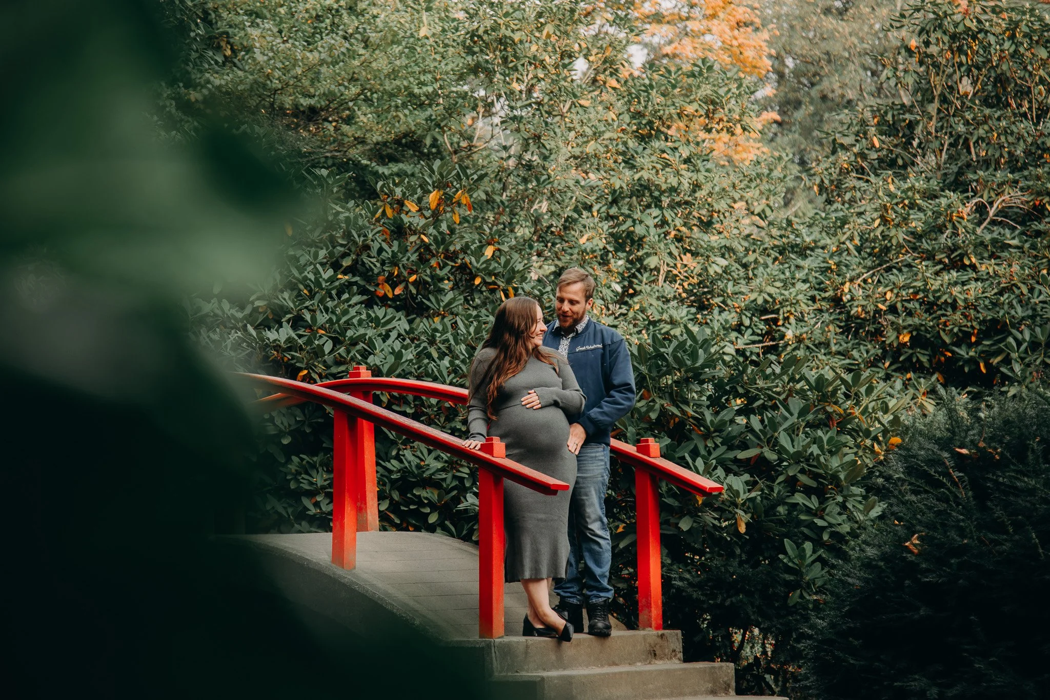 A couple stands on a small bridge surrounded by lush greenery, with the woman visibly pregnant. The woman is pregnant and wears a gray dress and black heels, while the man wears a blue jacket and jeans, looking at her affectionately.