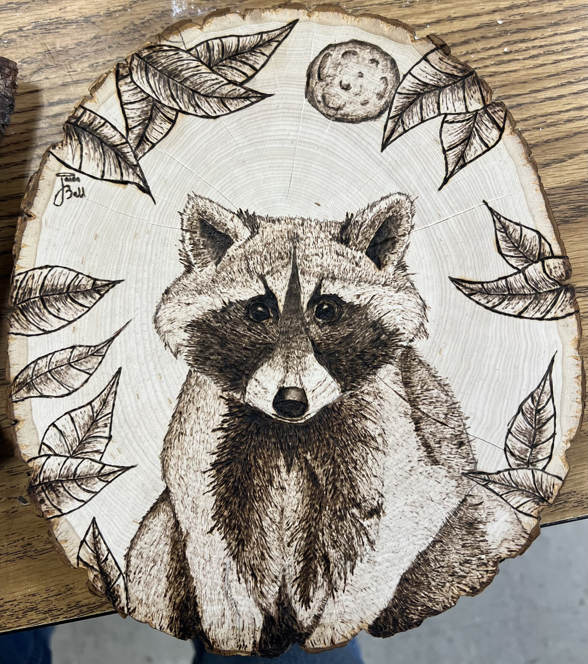 Burnt wood art of a raccoon's face on a tree slice, surrounded by engraved leaves and a moon with a face in the sky.