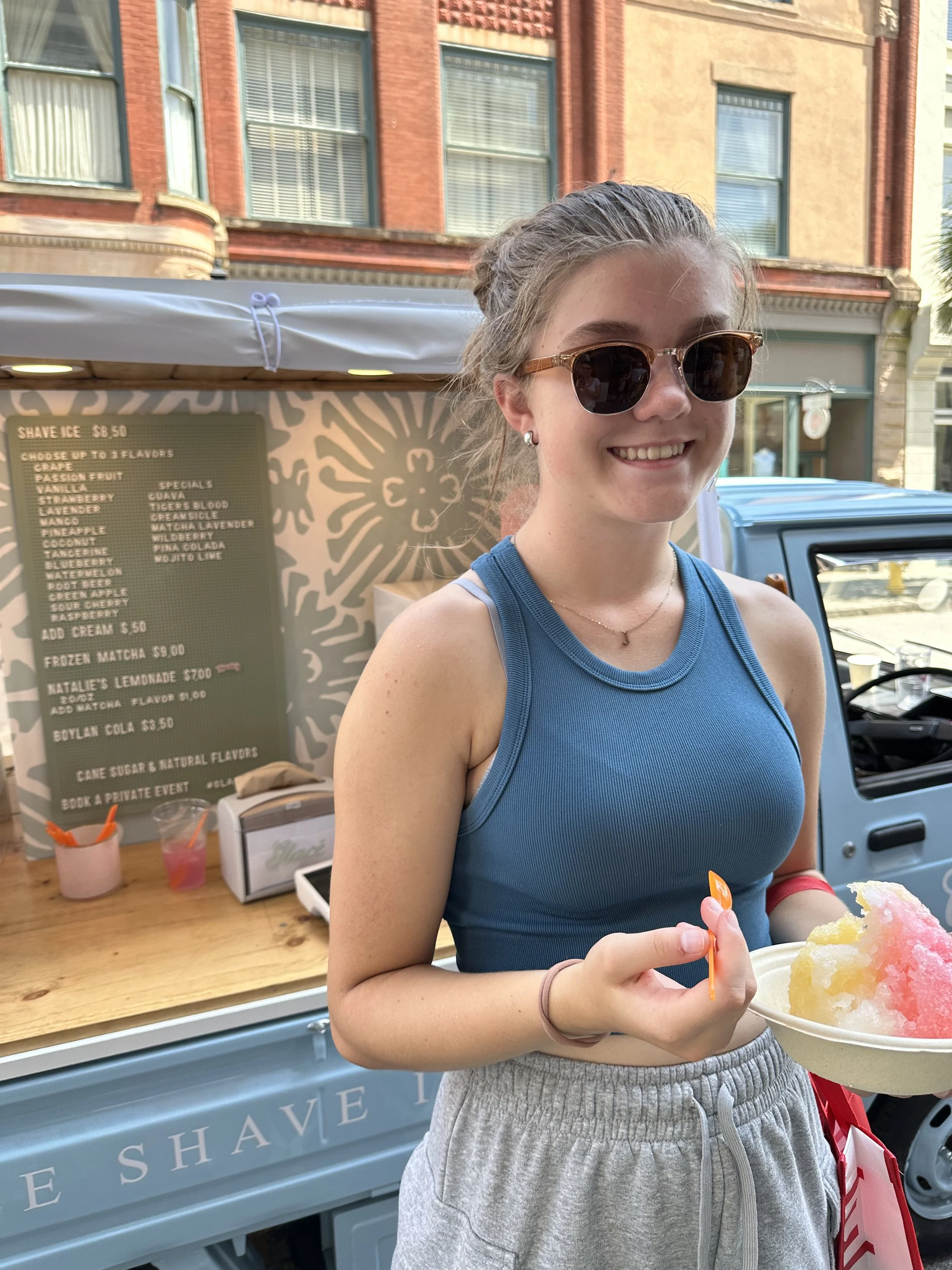 Young woman wearing sunglasses and a blue tank top holding a bowl of shaved ice with pink and yellow syrup, standing in front of a shaved ice stand.
