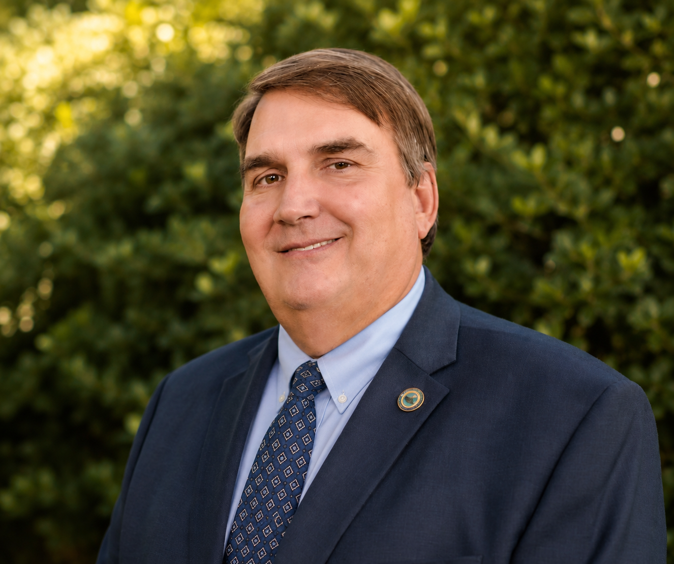 A portrait of a man in a navy suit with a blue tie, standing outdoors with greenery in the background. He is smiling and has a lapel pin on his suit.
