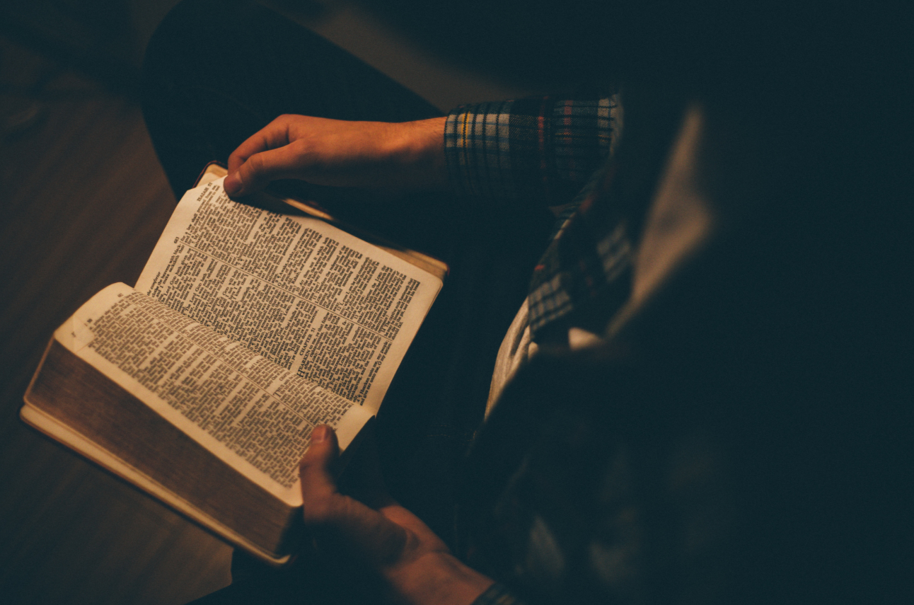 A person in a plaid sleeve reading a large, open book in a dimly lit room.