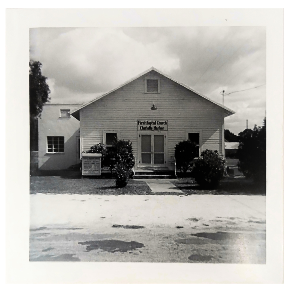 Black and white photo of a church building with a sign that reads "First Baptist Church Charlotte Harper" and some bushes in front.