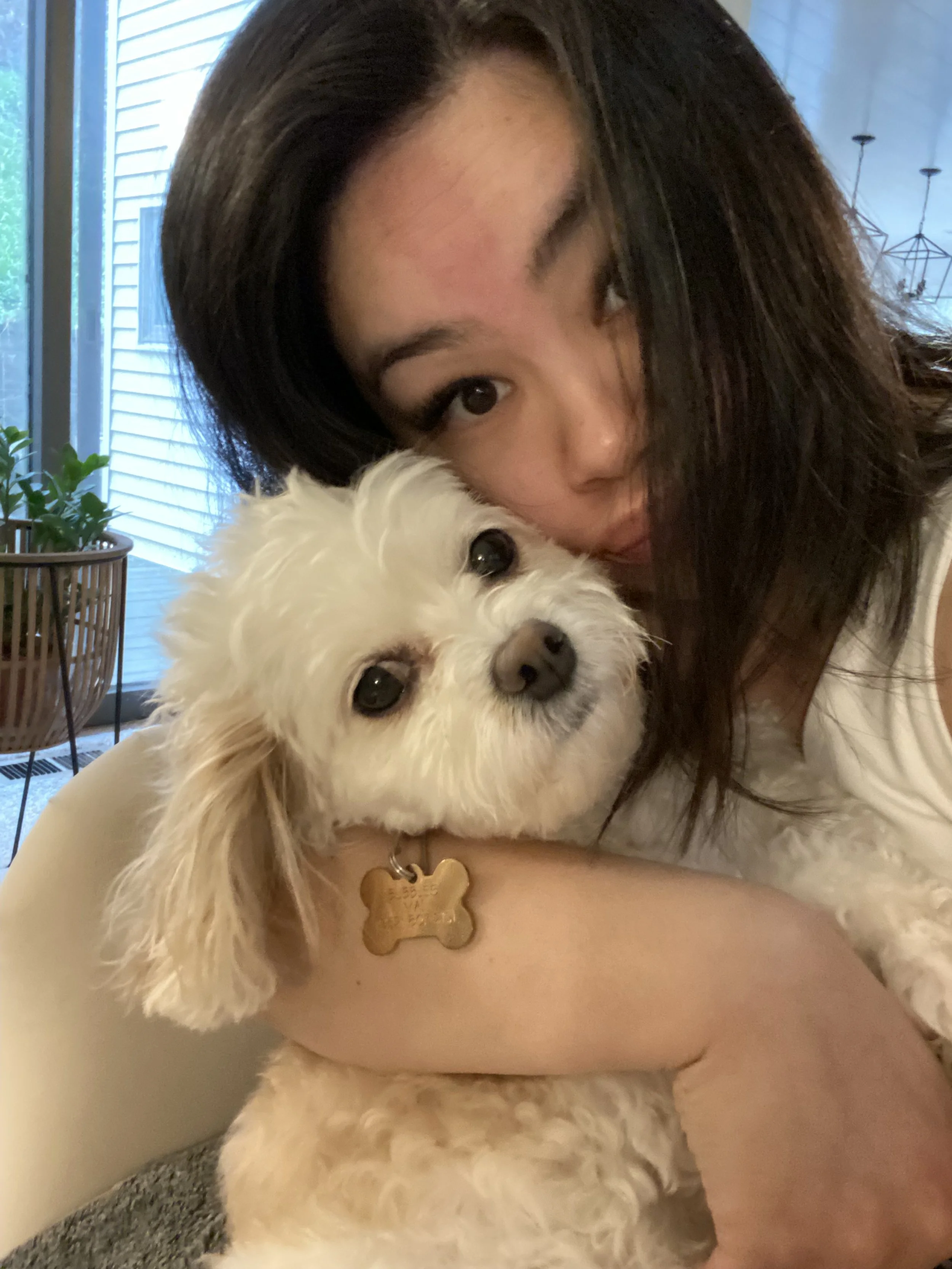A woman with long dark hair holding a small white dog with curly fur close to her face inside a home.