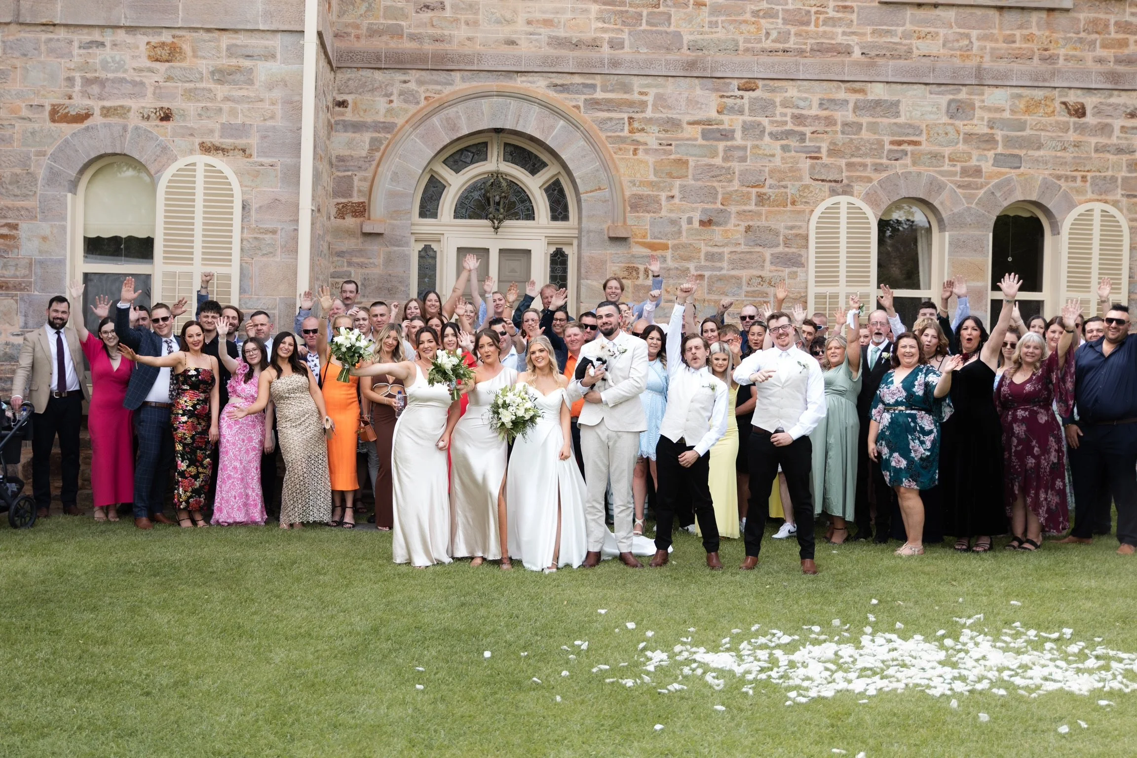 Large group of wedding guests and the bride and groom posing outside in front of a brick building, with some guests raising their hands in celebration.