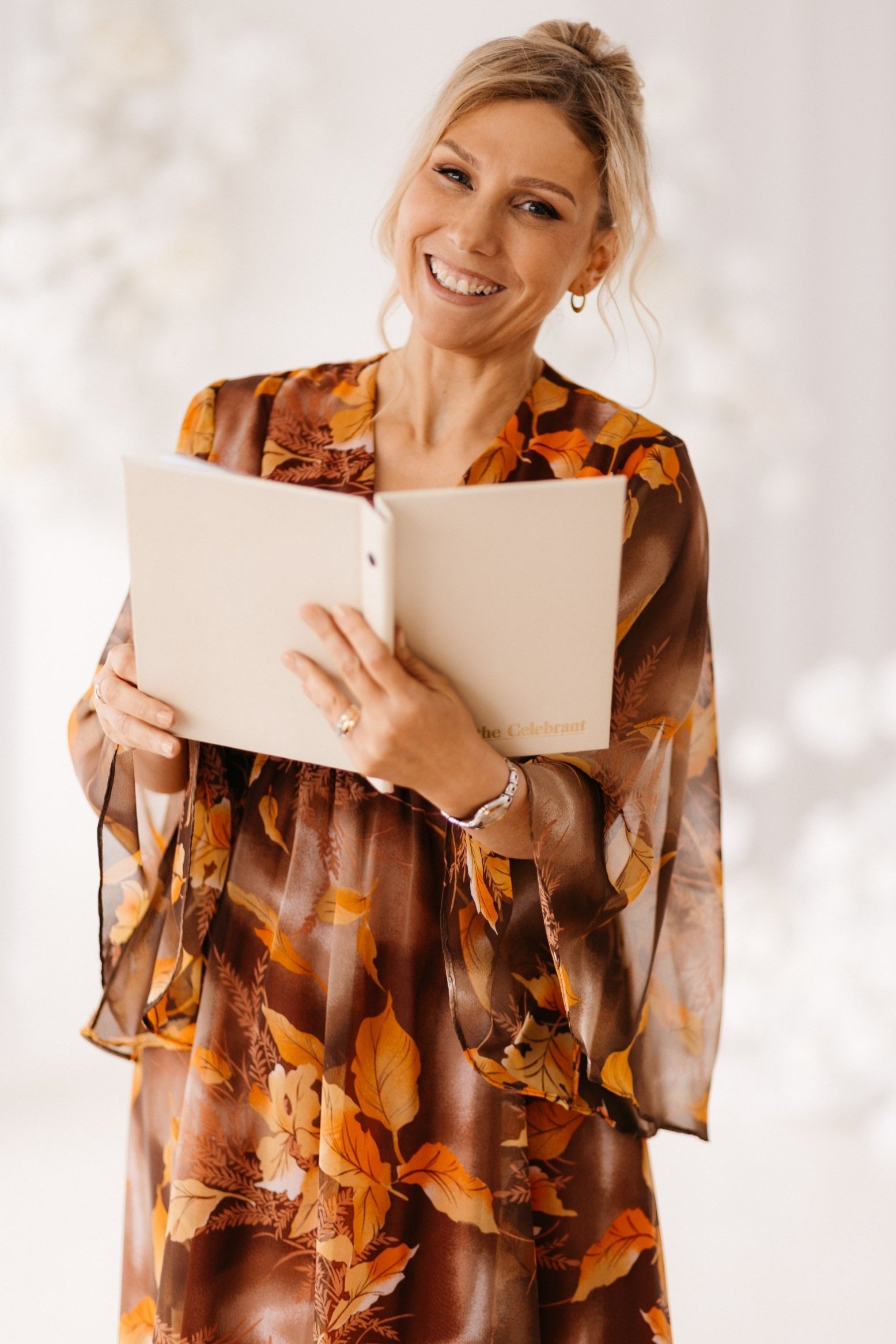 A woman smiling and holding an open book, dressed in a brown and orange floral patterned dress, with a blurred light-colored background.