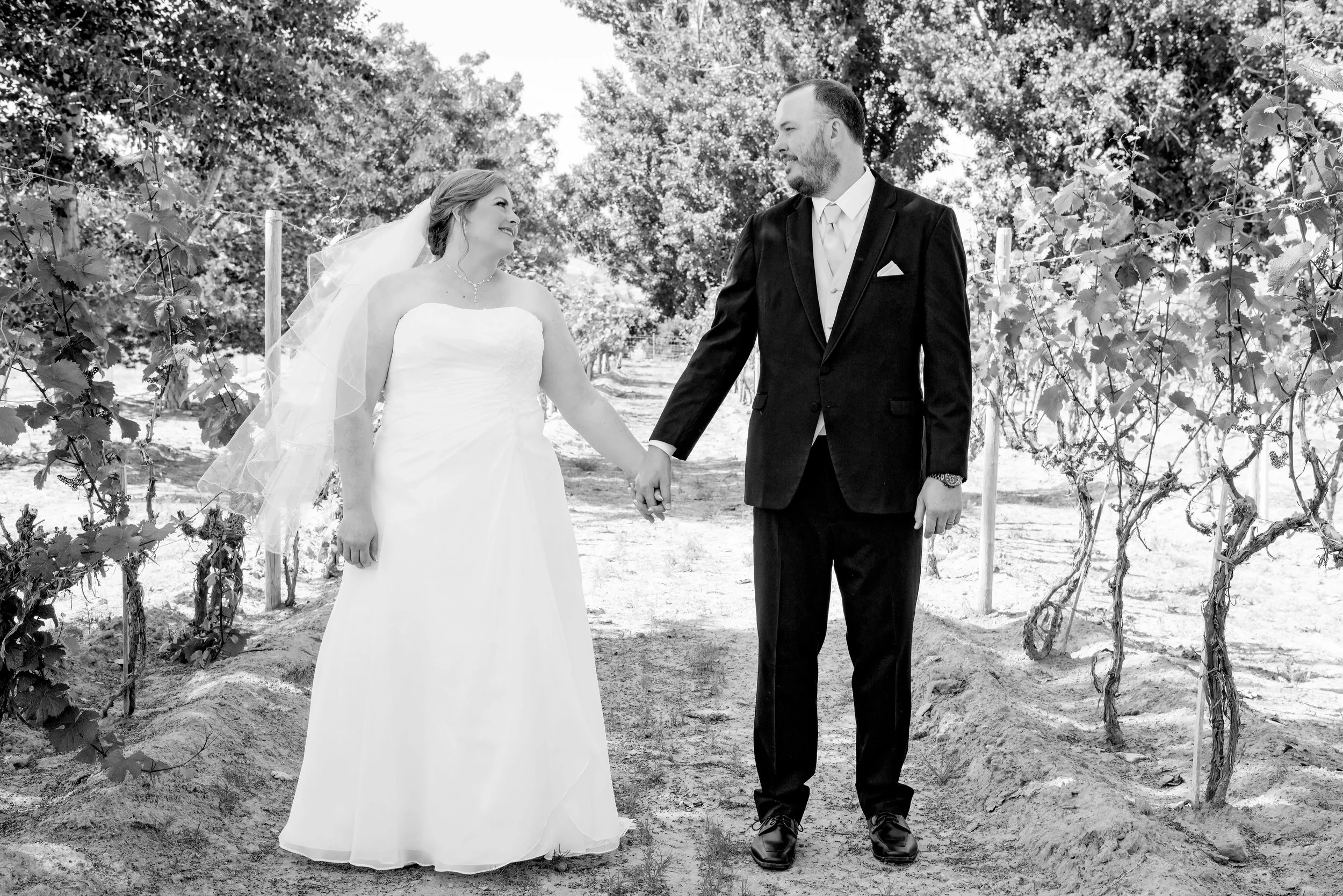 A bride and groom holding hands and smiling at each other in a vineyard, black and white photo.