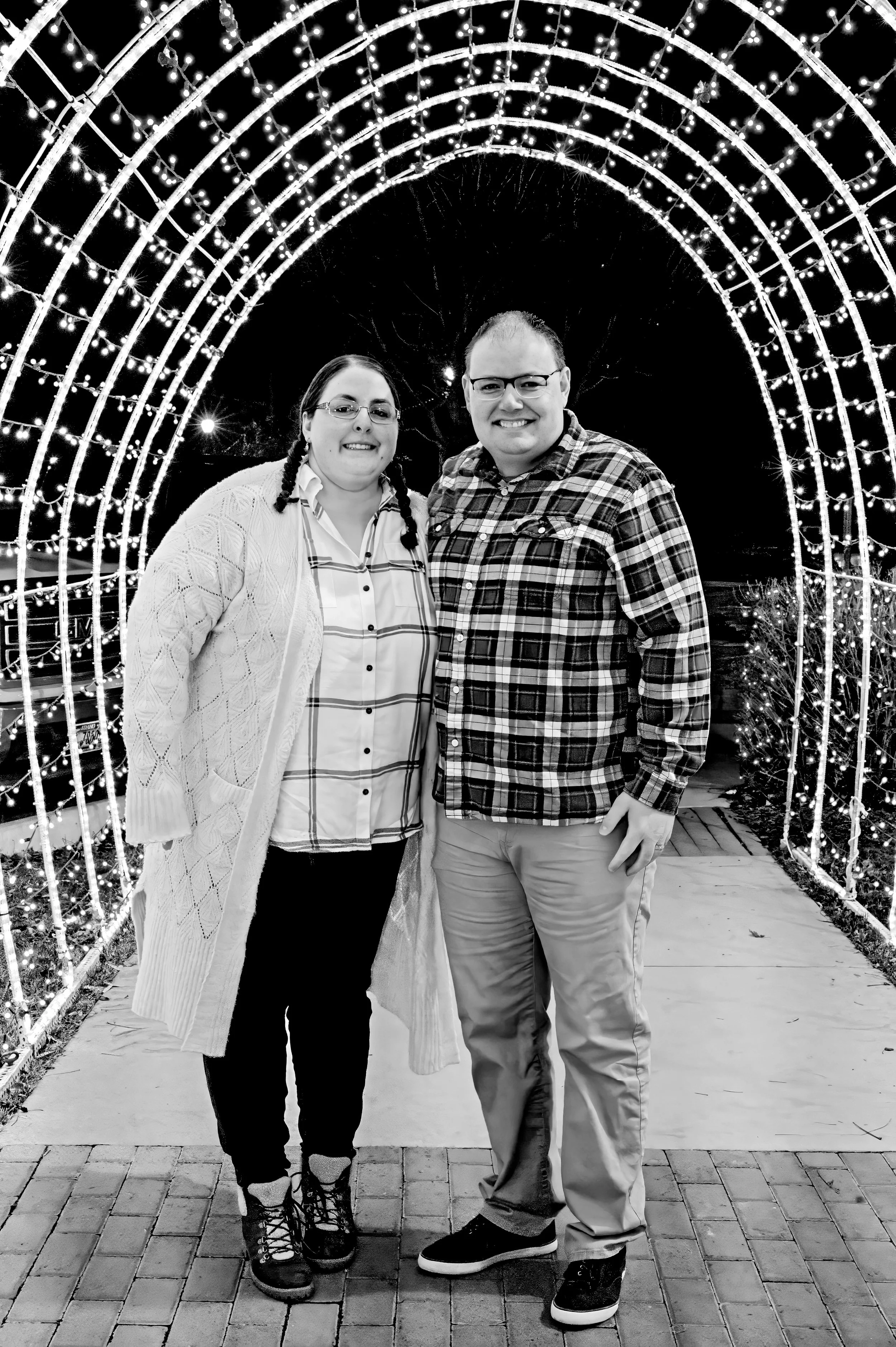 A smiling couple stands under a decorated archway of holiday lights at night, with trees and lights in the background.
