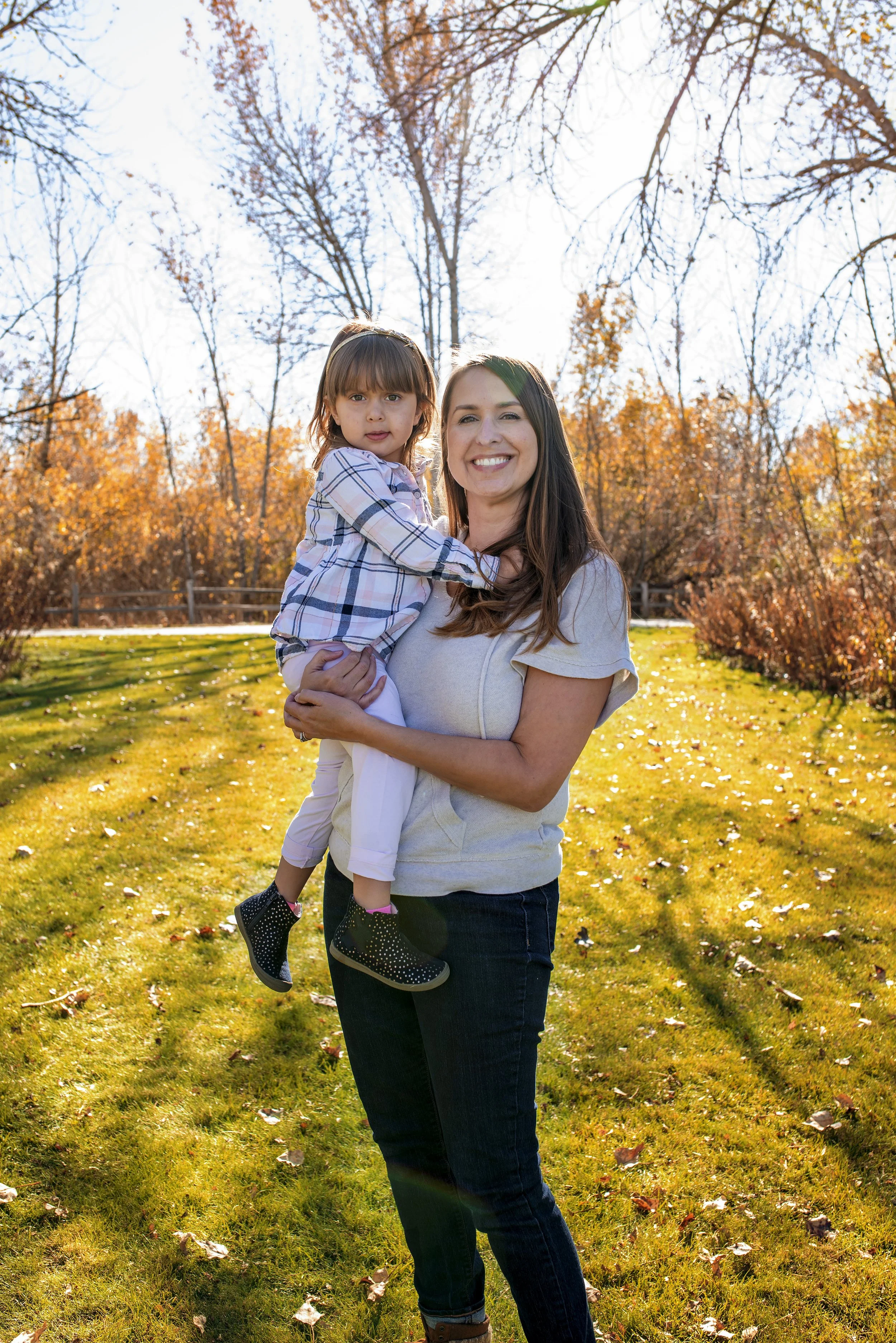 A woman holding a young girl outdoors on a sunny autumn day, with trees with orange leaves in the background and a grassy area.