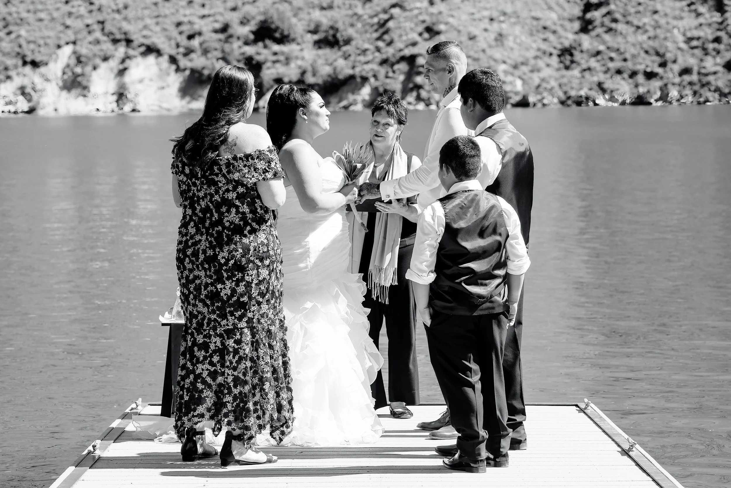 A wedding ceremony taking place on a dock beside a body of water with six people standing in a line, including the bride, groom, officiant, and three witnesses or family members.