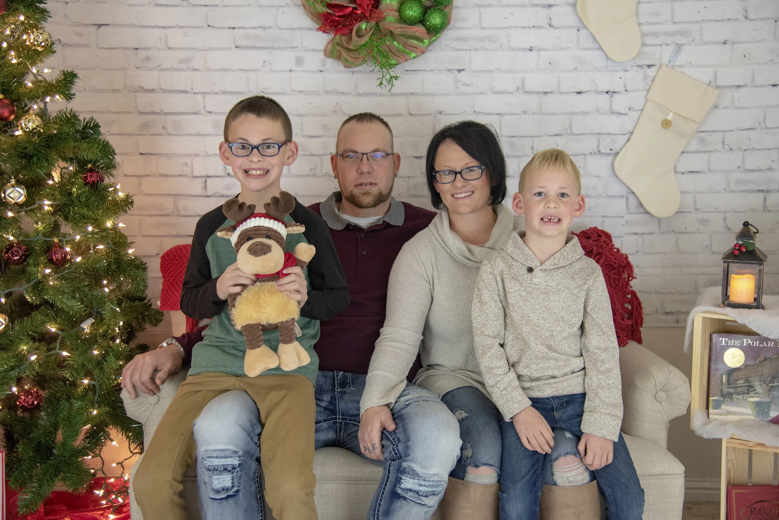 A family sitting on a beige couch in a Christmas decorated room, with a Christmas tree, stockings, and holiday decorations on the wall.