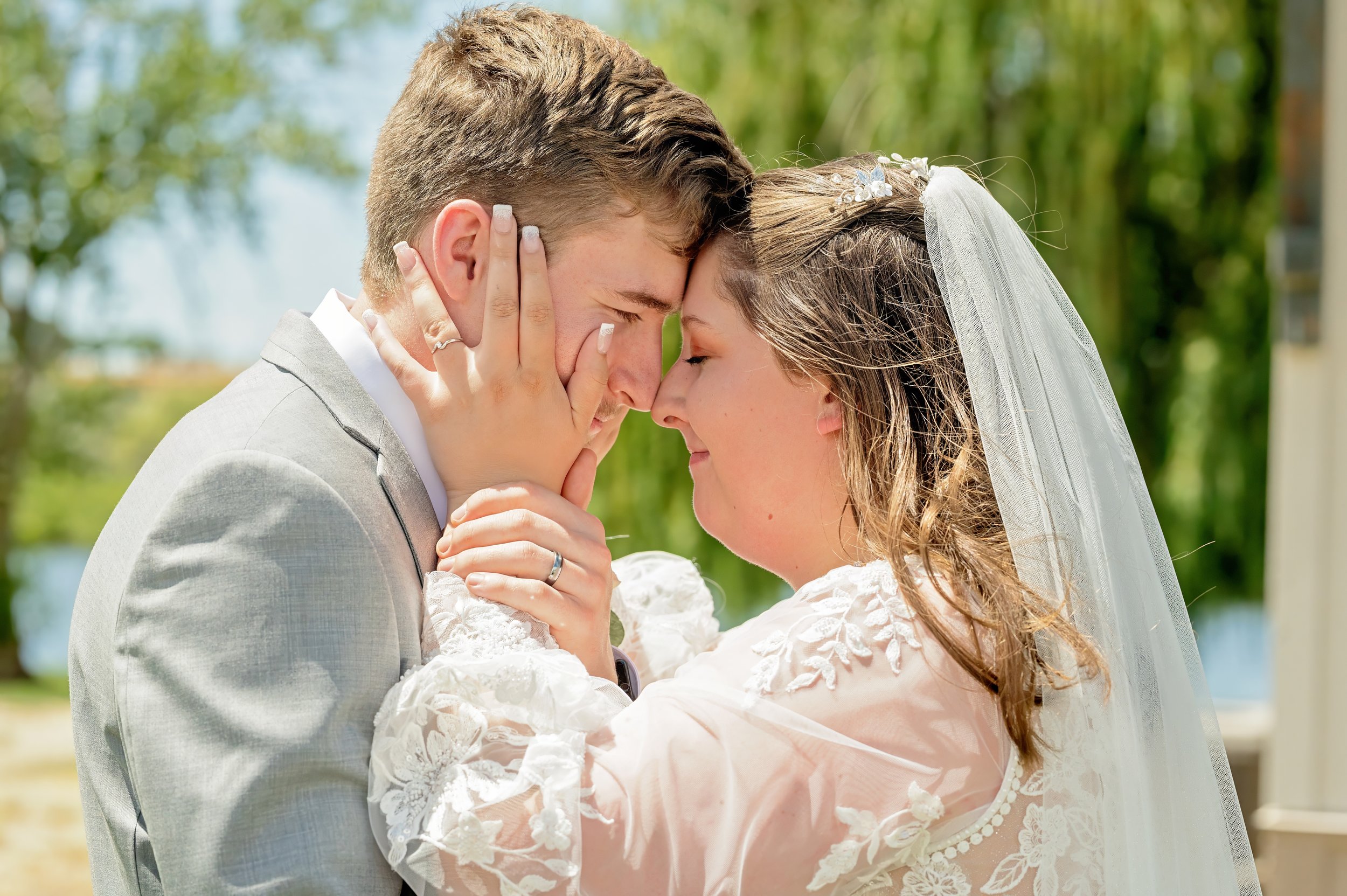 A bride and groom are touching foreheads outdoors on a sunny day, with greenery and water in the background.