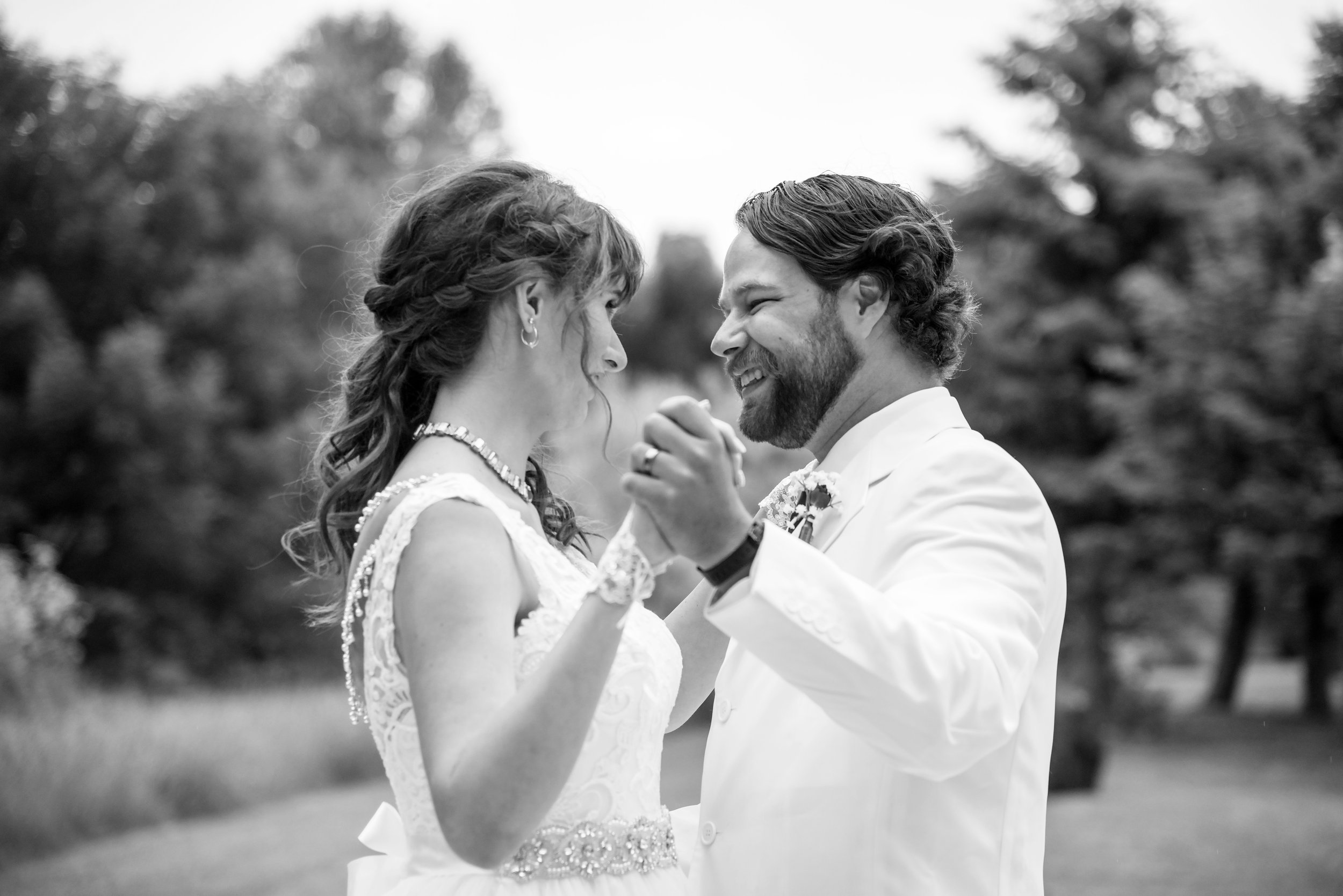 A black-and-white photo of a bride and groom dancing outdoors, smiling and looking into each other's eyes.