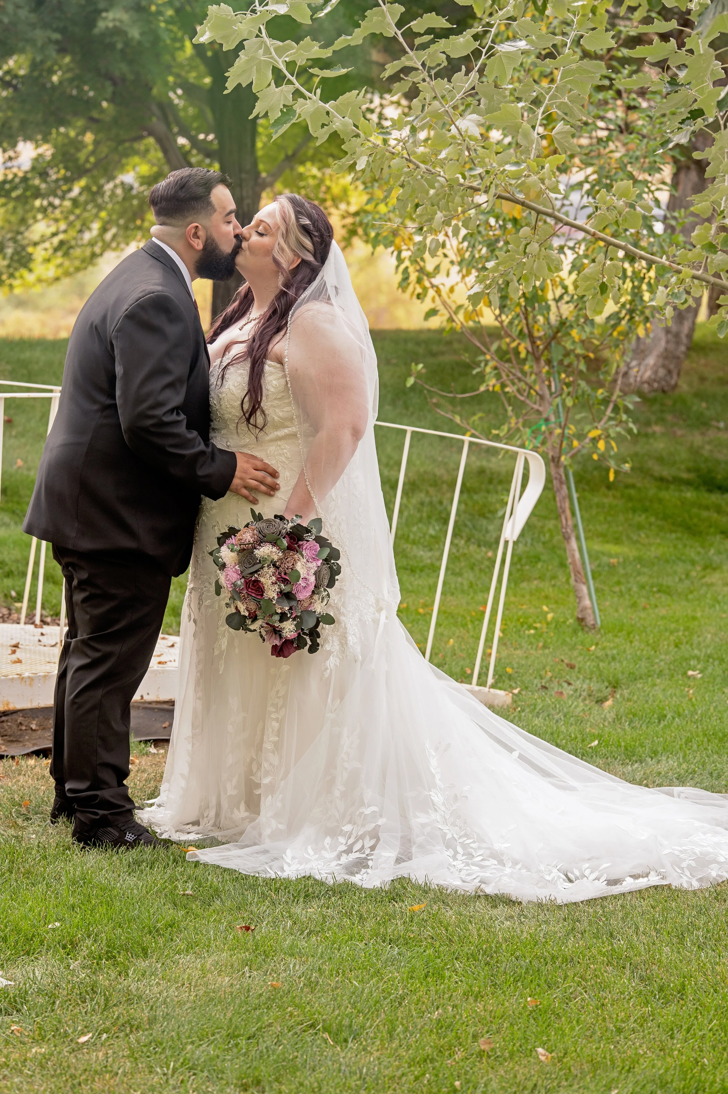 A bride and groom sharing a kiss outdoors on their wedding day, with the bride holding a bouquet of pink, purple, and white flowers, standing on a grassy area with trees in the background.