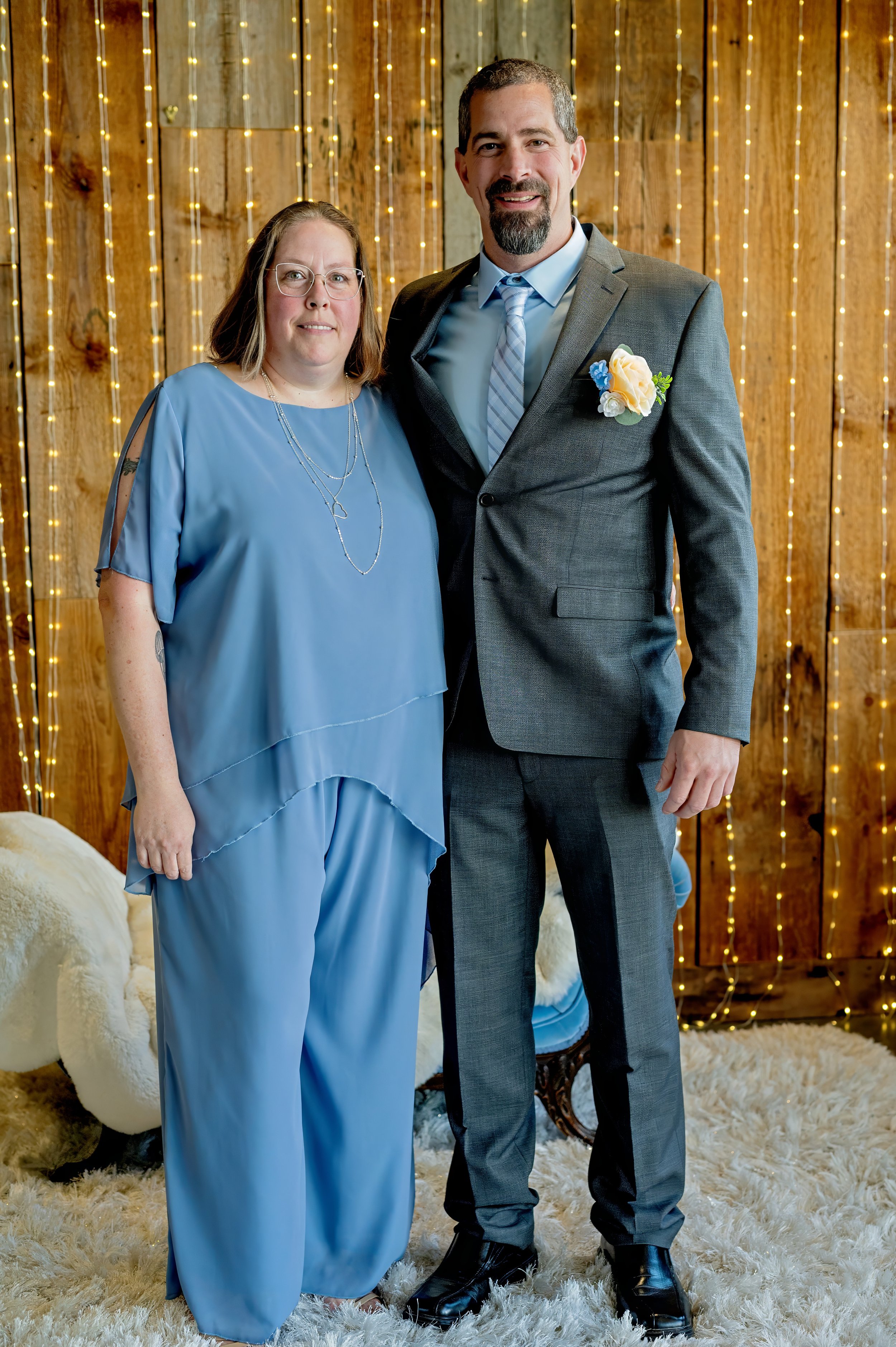 A woman and a man stand side by side indoors with a wooden wall decorated with string lights in the background. The woman wears a blue outfit with layered jewelry and glasses. The man is dressed in a gray suit with a light blue shirt, striped tie, and a boutonniere pinned to his lapel.