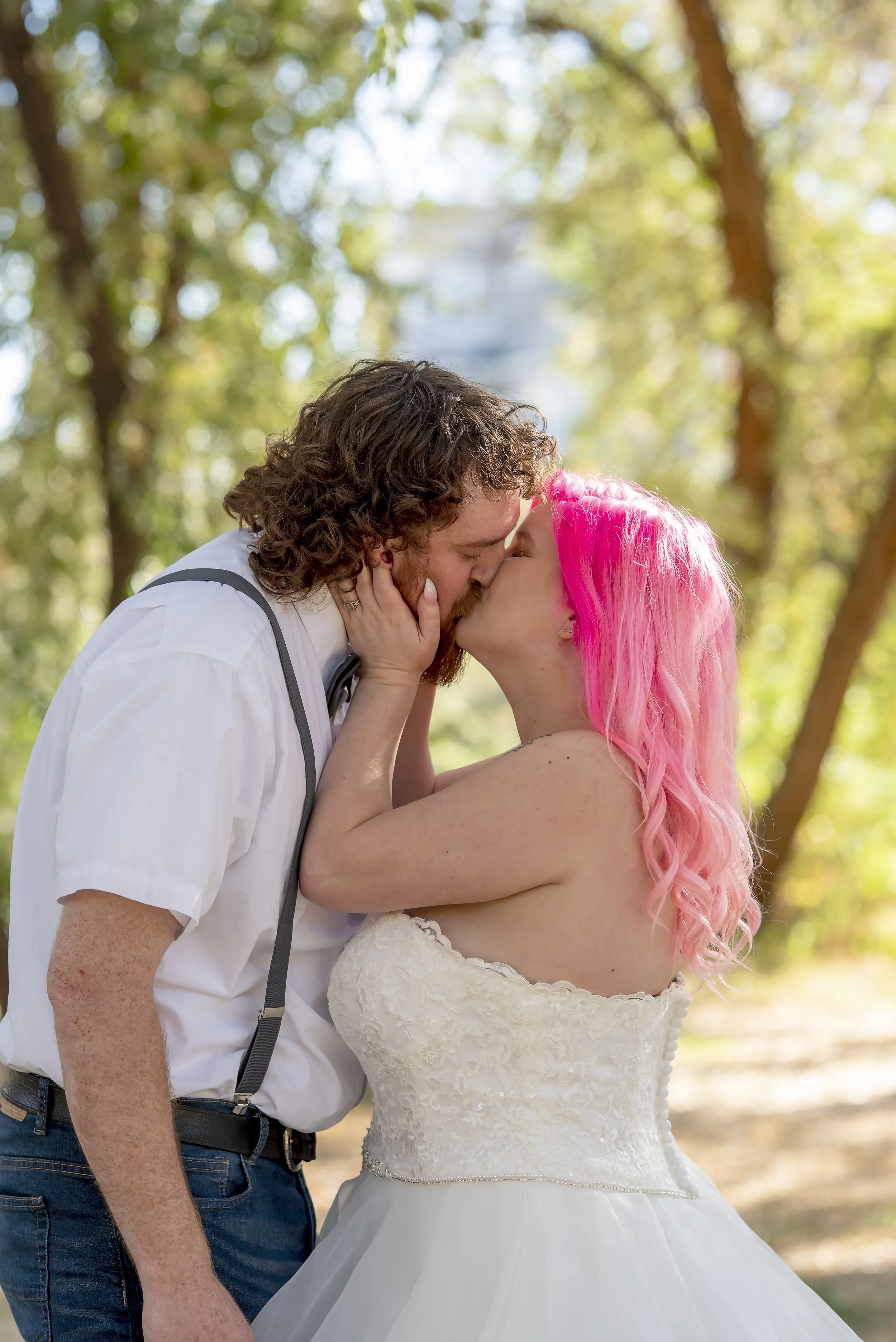 A couple kissing outdoors, with the woman having pink hair and the man wearing a white shirt and suspenders, in a forest setting.