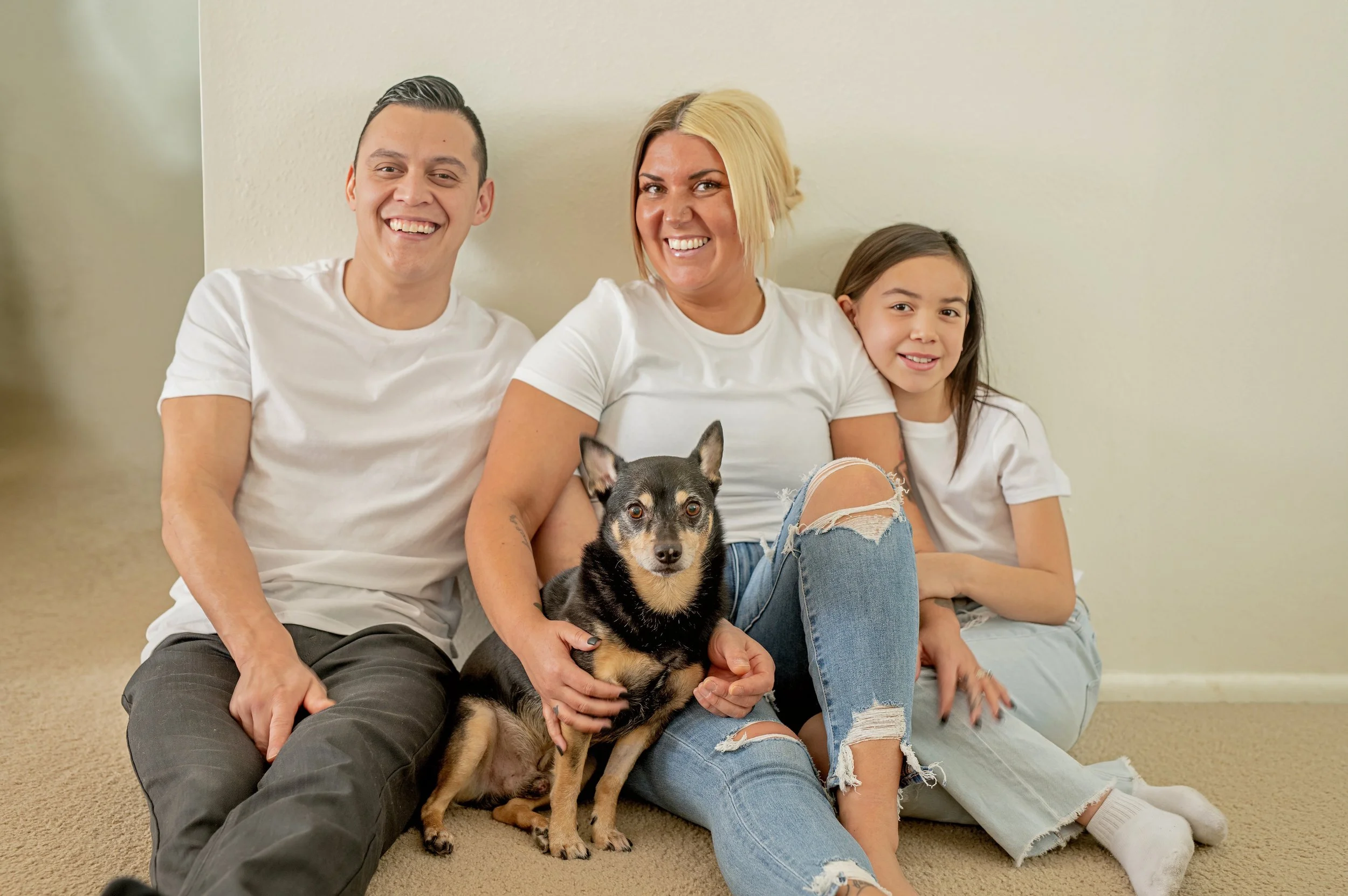 A smiling family of three and a dog sitting on a carpeted floor against a wall, all wearing white shirts.