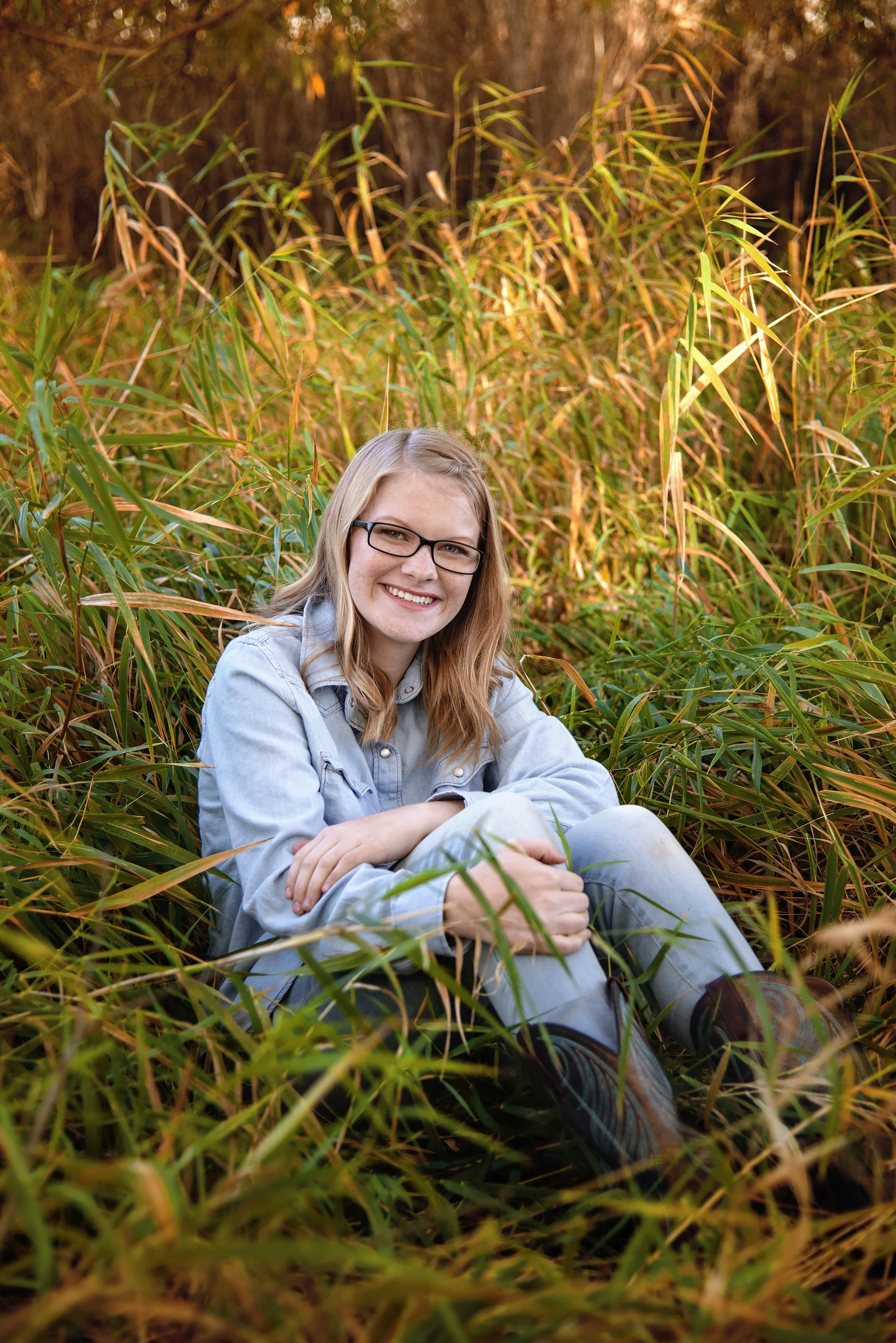 A young woman with glasses and long blonde hair, wearing a light blue denim jacket and jeans, sitting among tall, golden-brown grass and green plants outdoors, smiling at the camera.