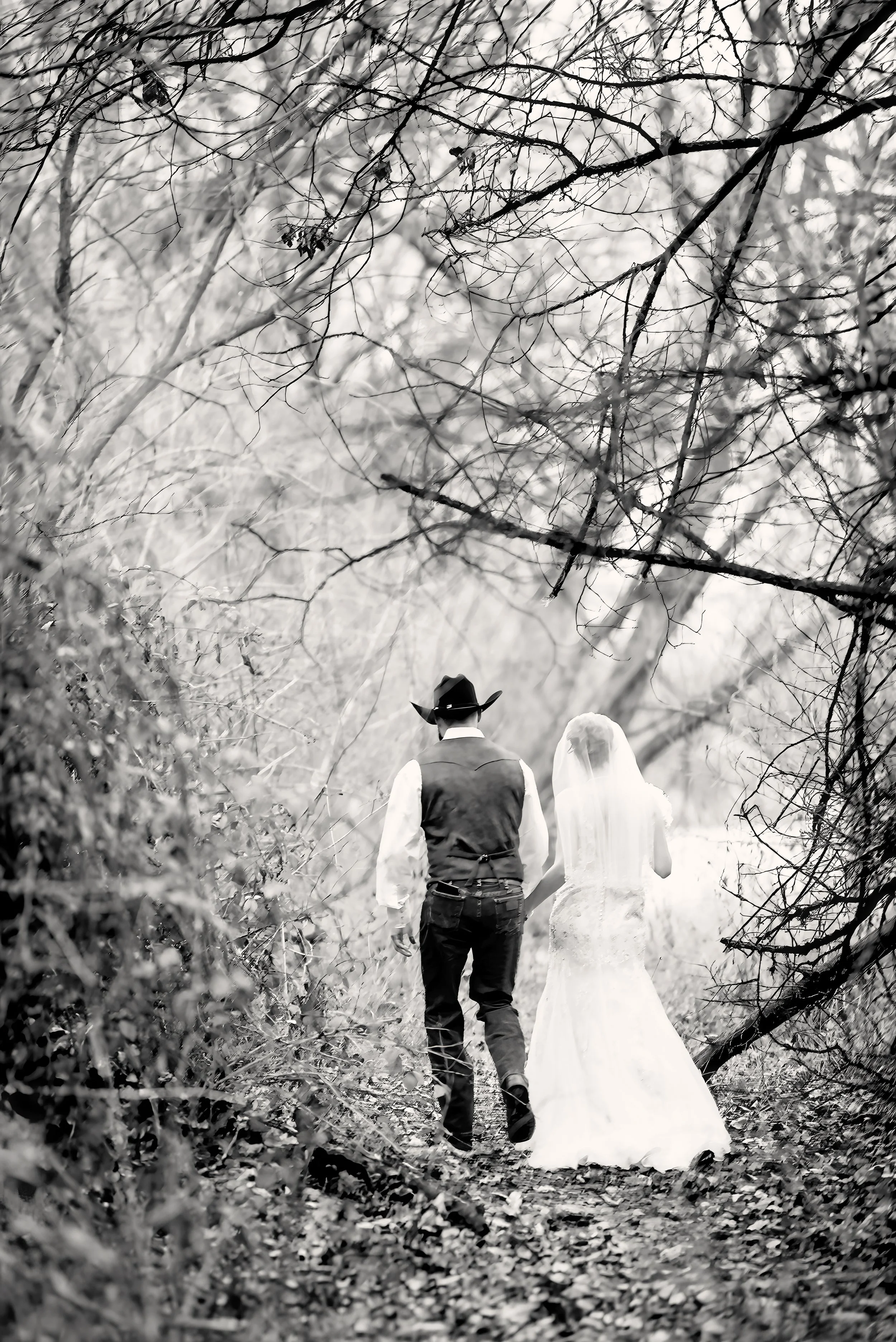 A bride and groom walk hand-in-hand through a wooded area, with leafless branches overhead, in black and white.