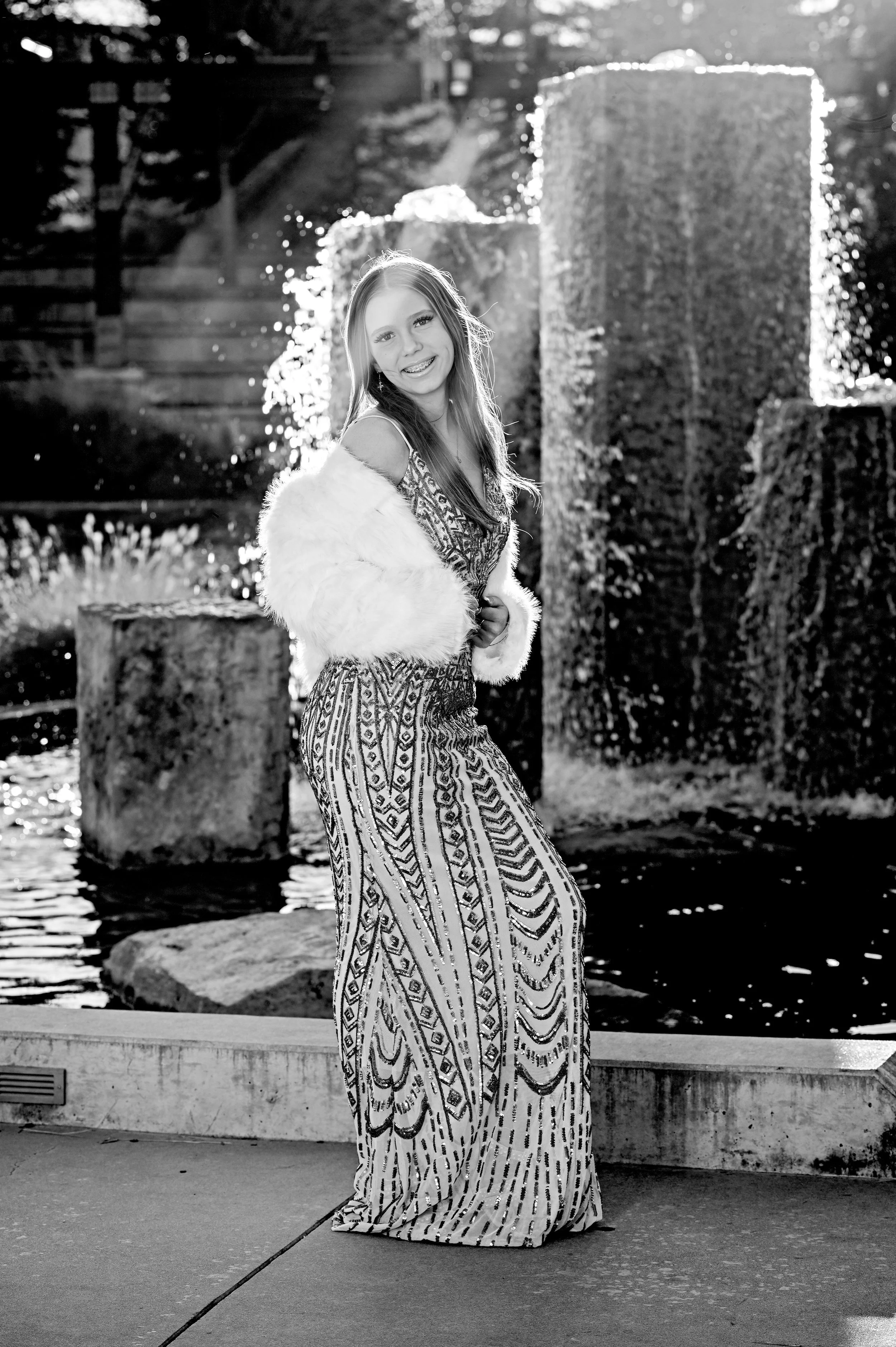 A young girl smiling and posing next to a water fountain with cascading water, wearing a patterned dress and a fluffy jacket.