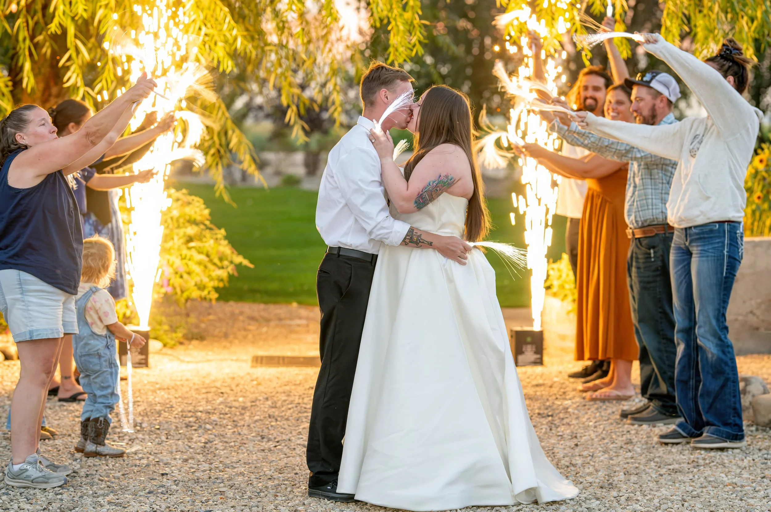 A newlywed couple shares a kiss during their outdoor wedding celebration at sunset, surrounded by friends and family holding sparklers and celebrating.