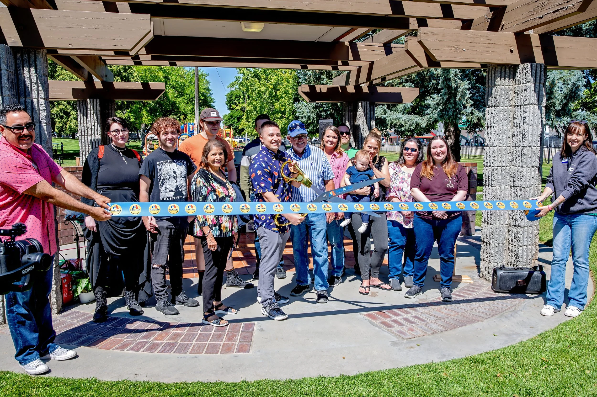 Group of people at a ribbon-cutting ceremony outdoors, holding a ribbon with a scissors and a leaf at the center, under a wooden and stone structure, with trees and a playground in the background.