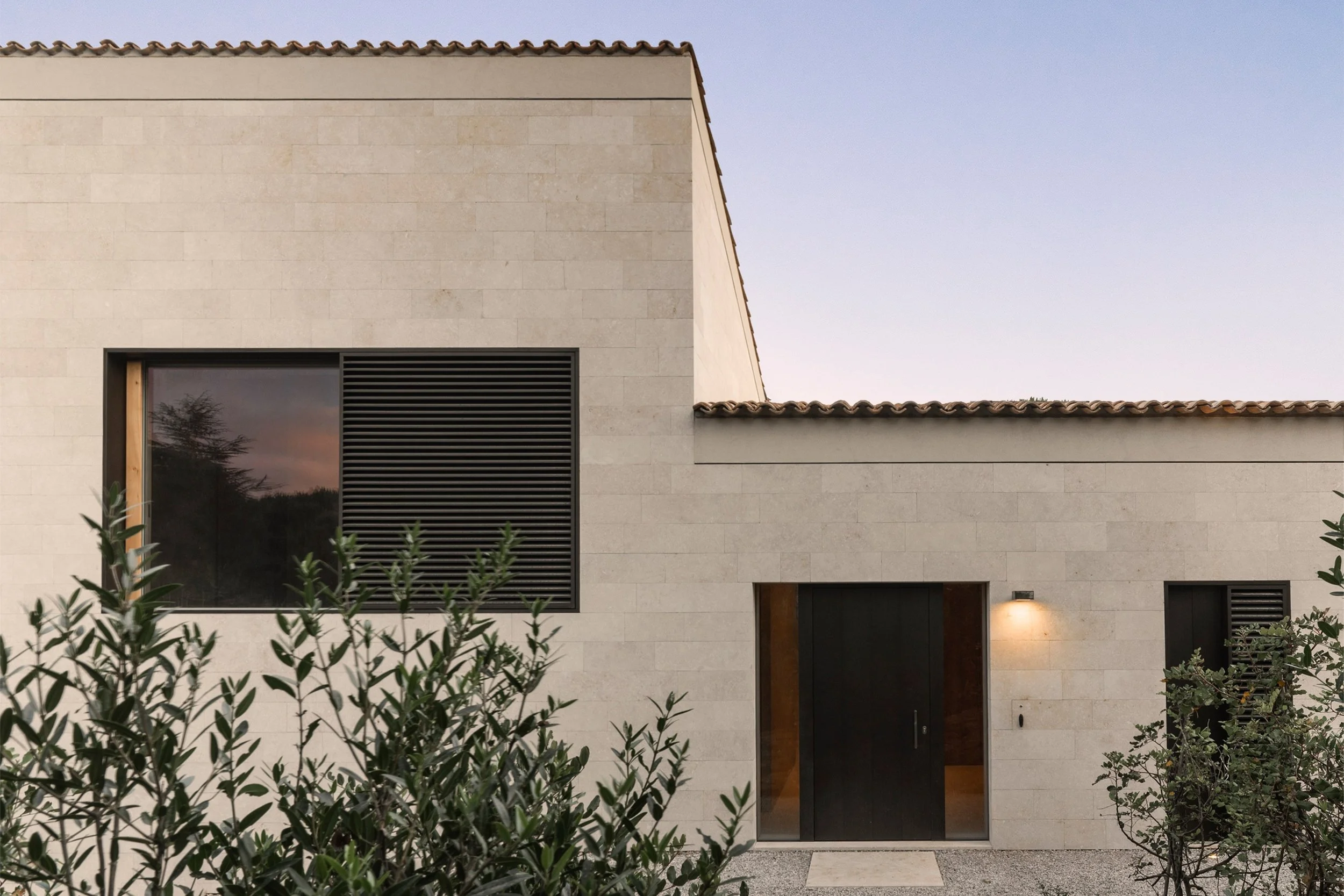 Modern beige house exterior with large window, black door, and outdoor lighting, surrounded by green bushes during dusk or dawn.