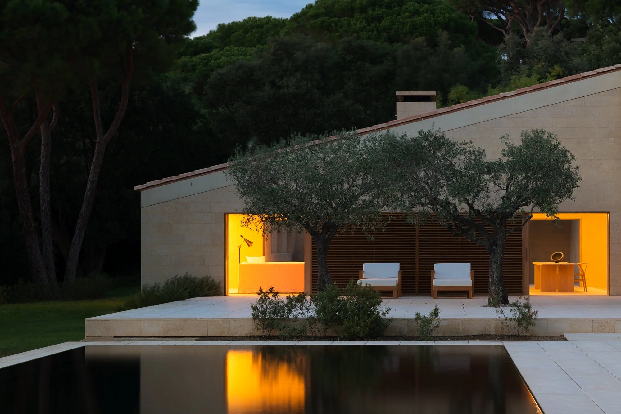 Modern house exterior with two trees in front, illuminated yellow interior rooms, patio seating, and a reflecting pool in the foreground, surrounded by greenery.