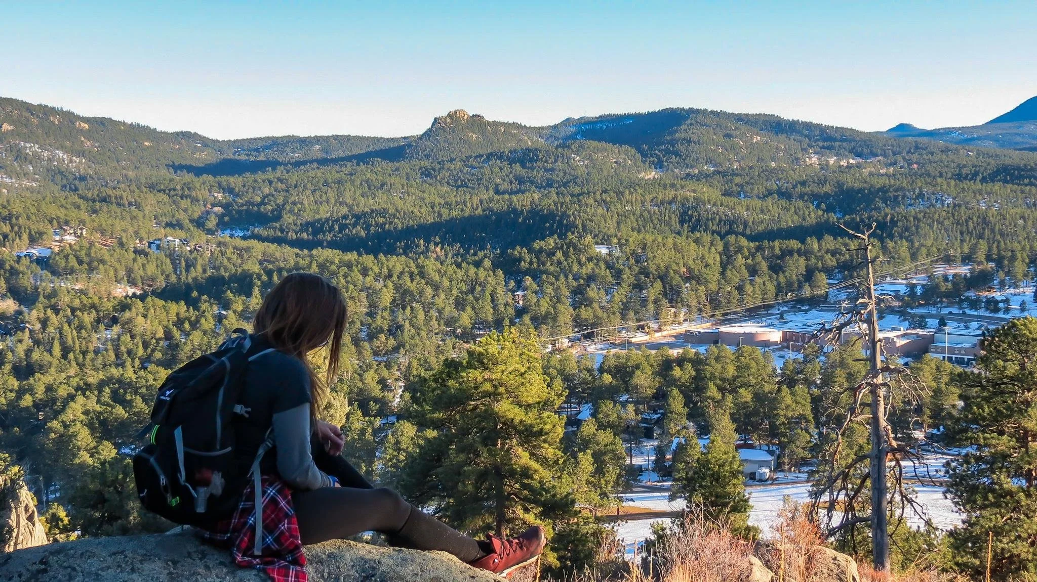 A woman sitting on a rock overlooking a sprawling forested valley with mountains in the background, some areas covered in snow, under a clear blue sky.