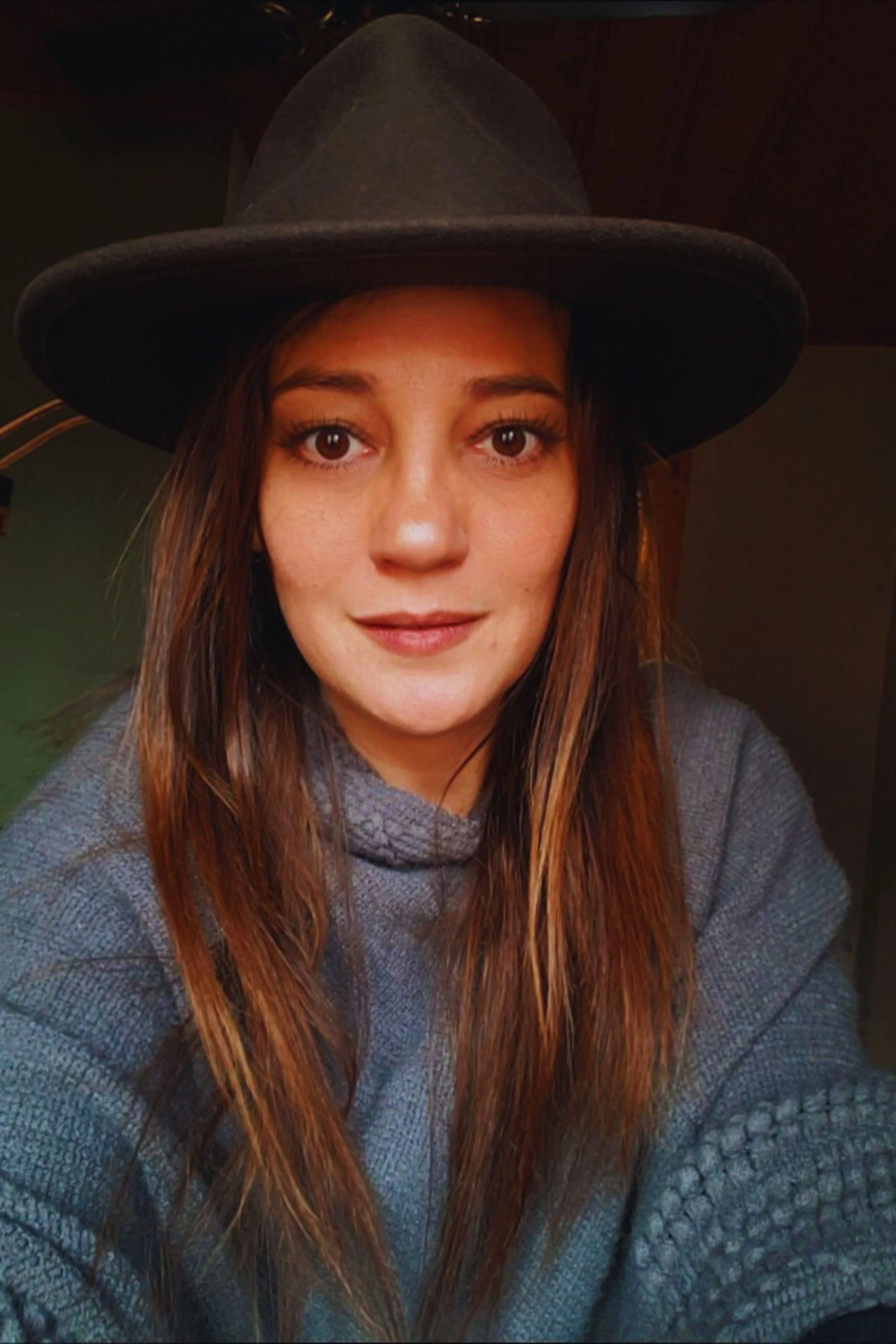 A woman with long, light brown hair wearing a gray sweater and a large black hat, smiling at the camera indoors.